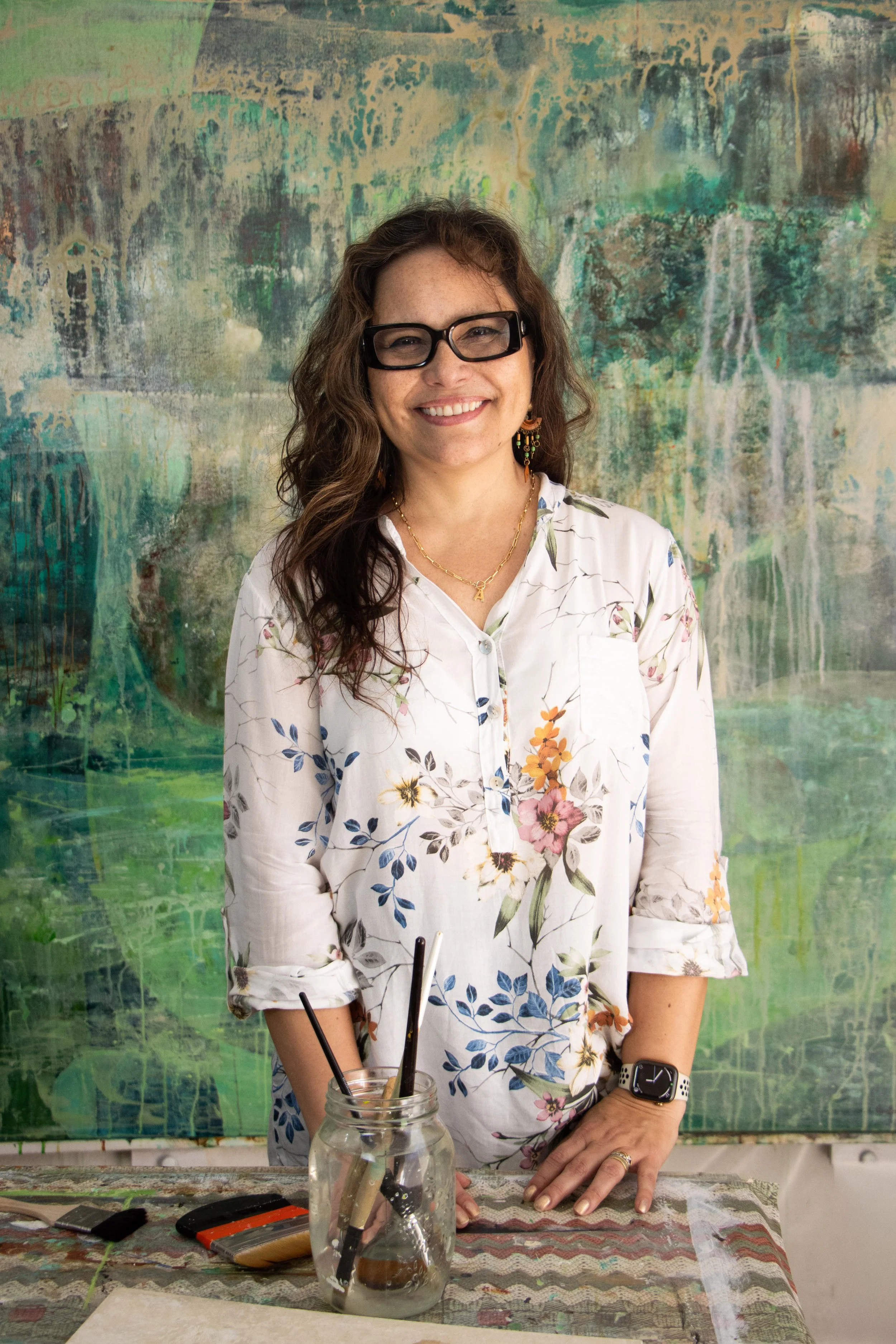 A smiling woman with wavy brown hair wearing glasses, a floral white shirt, and a necklace, standing in front of an abstract green and blue painting. She has one hand on a table with art supplies and objects, including paintbrushes and a mason jar filled with water.