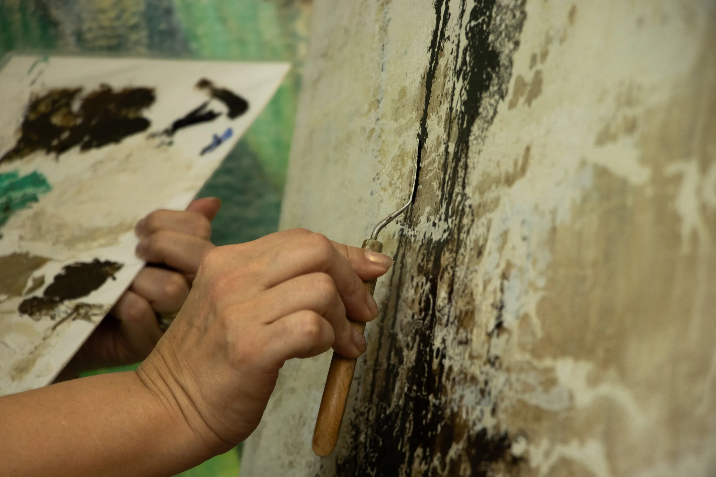 Close-up of a person using a small chisel and hammer to peel blackened material from a textured, aged wall.