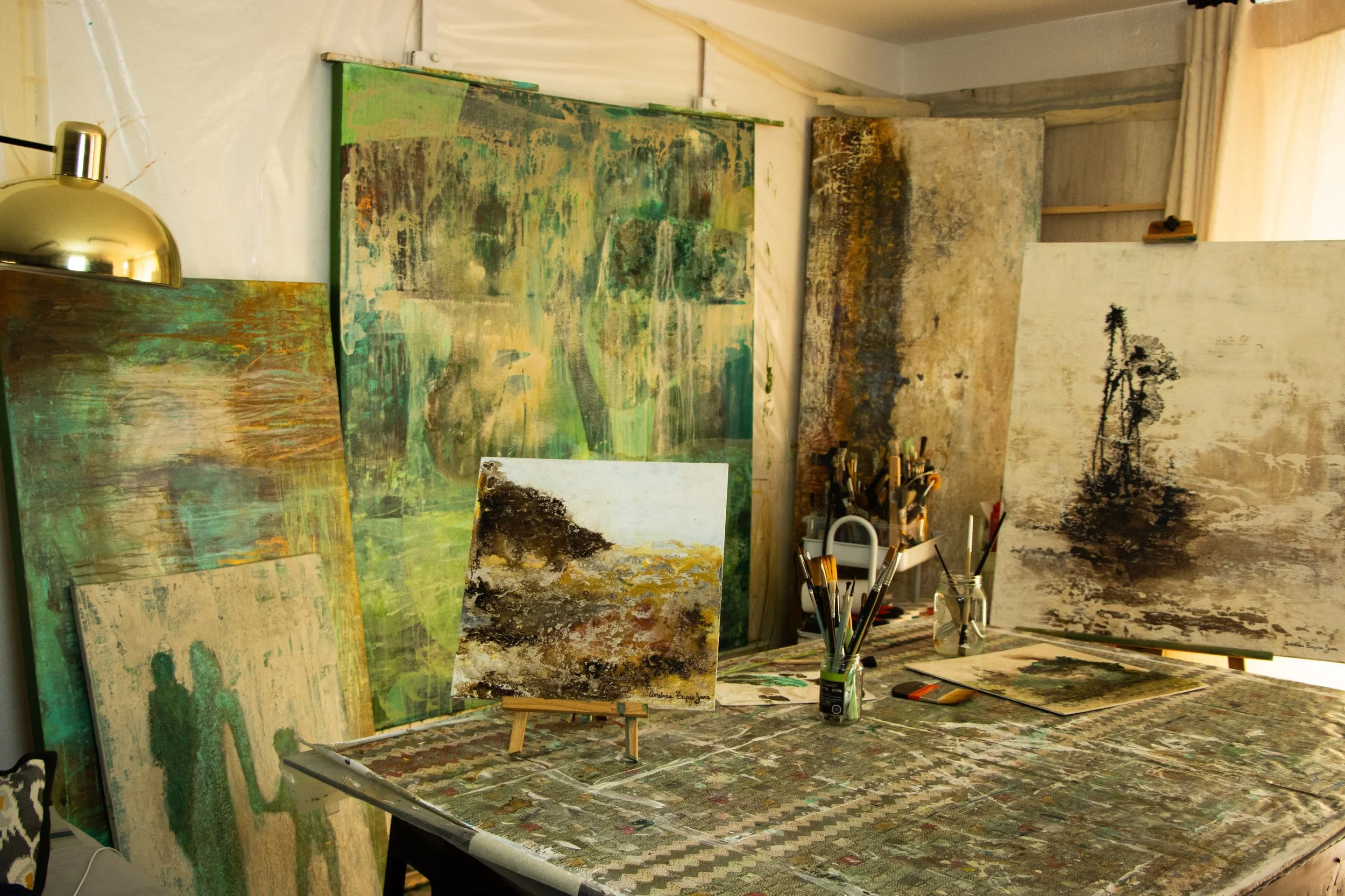 Art studio with multiple abstract landscape paintings leaning against the wall, some on easels, with brushes and painting supplies on the table and in jars.