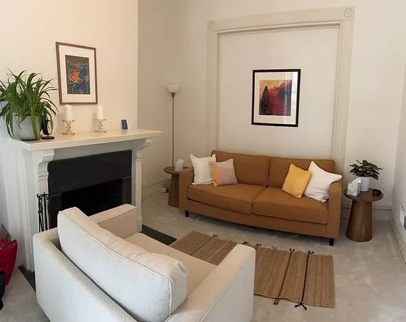 Living room with a brown couch with white and yellow pillows, beige armchair, fireplace, framed artwork, and a small wooden side table with a plant.
