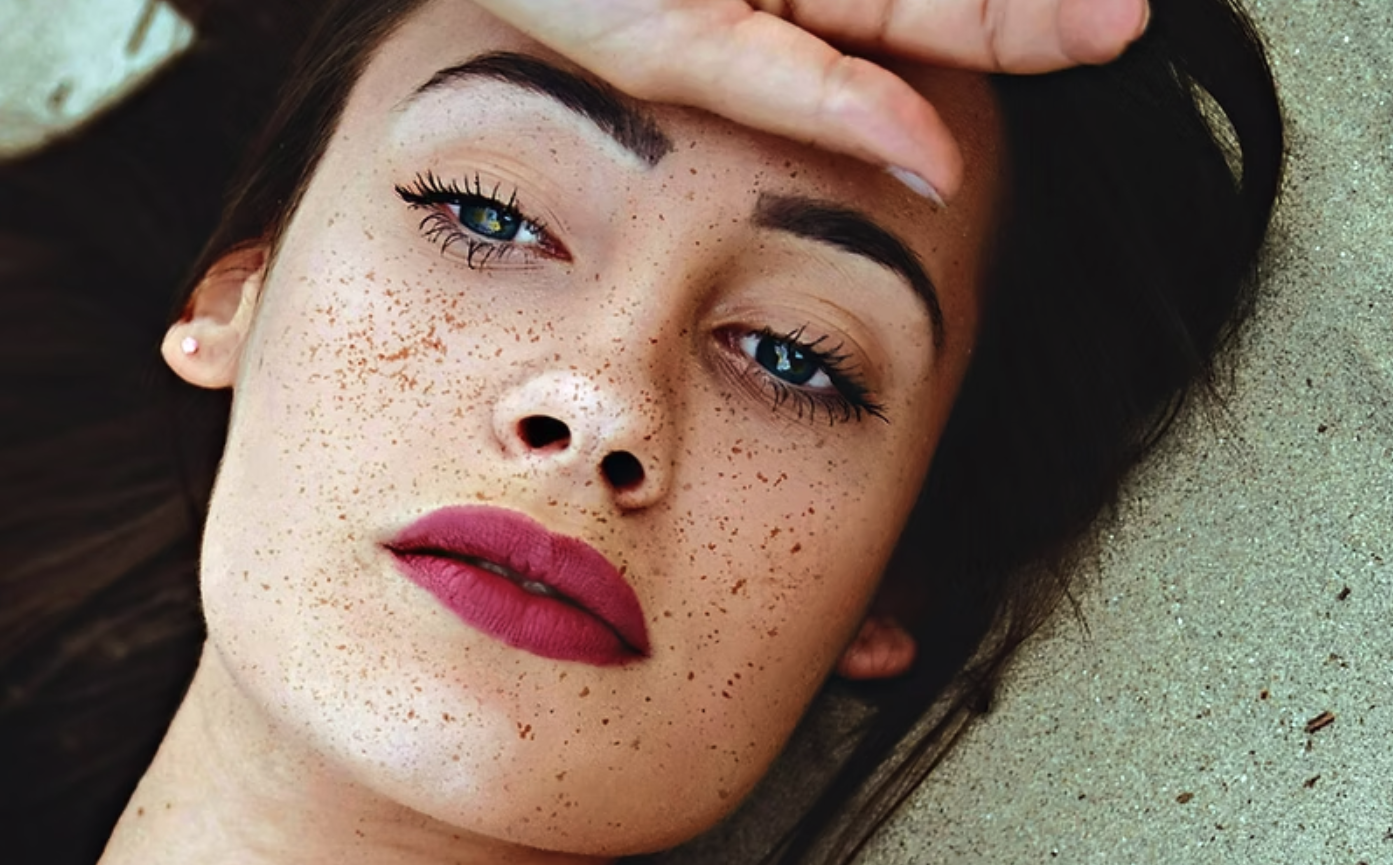 Close-up of a young woman with dark hair lying on sand, with freckles, bright blue eyes, dark eyebrows, pink lipstick, and a small pearl earring.