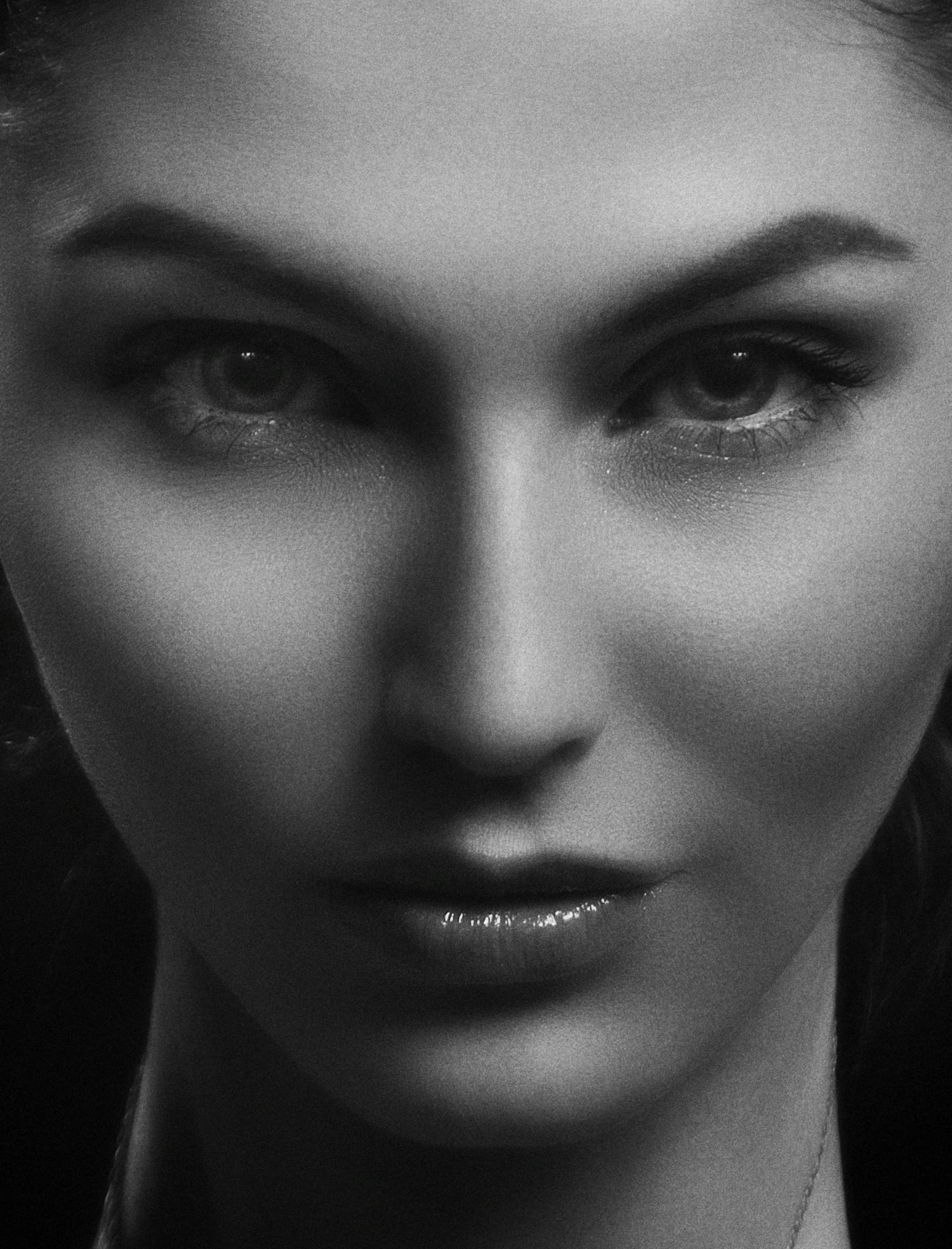 A close-up black and white photograph of a woman's face, focusing on her eyes, eyebrows, and part of her nose and lips, with the woman looking down slightly.