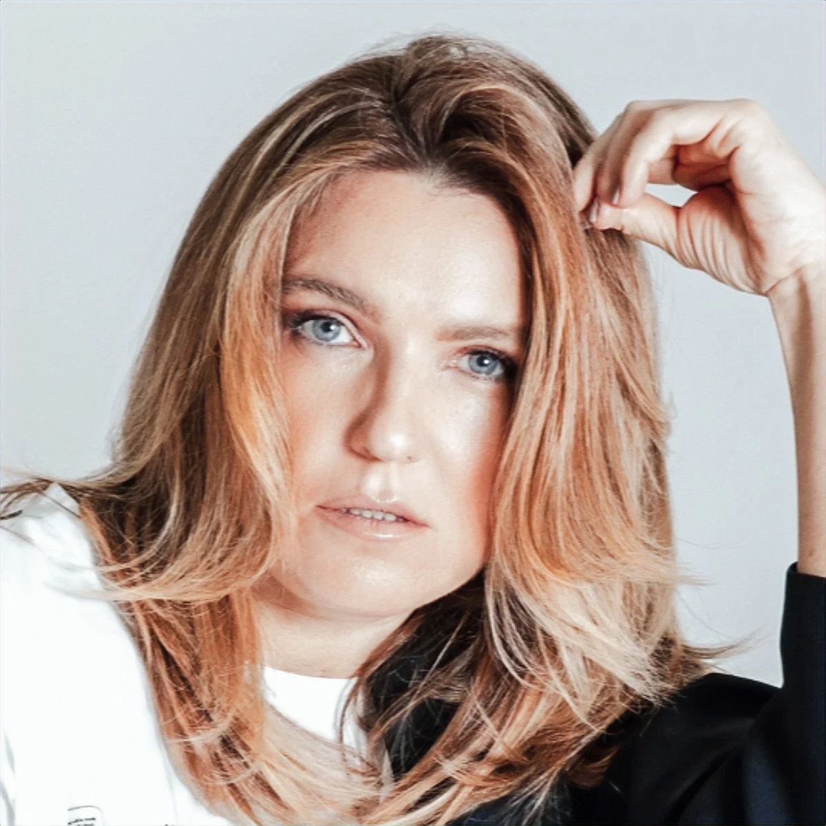 Close-up of Alisa Gualico  with wavy, light brown hair and blue eyes, looking at the camera with her hand touching her hair