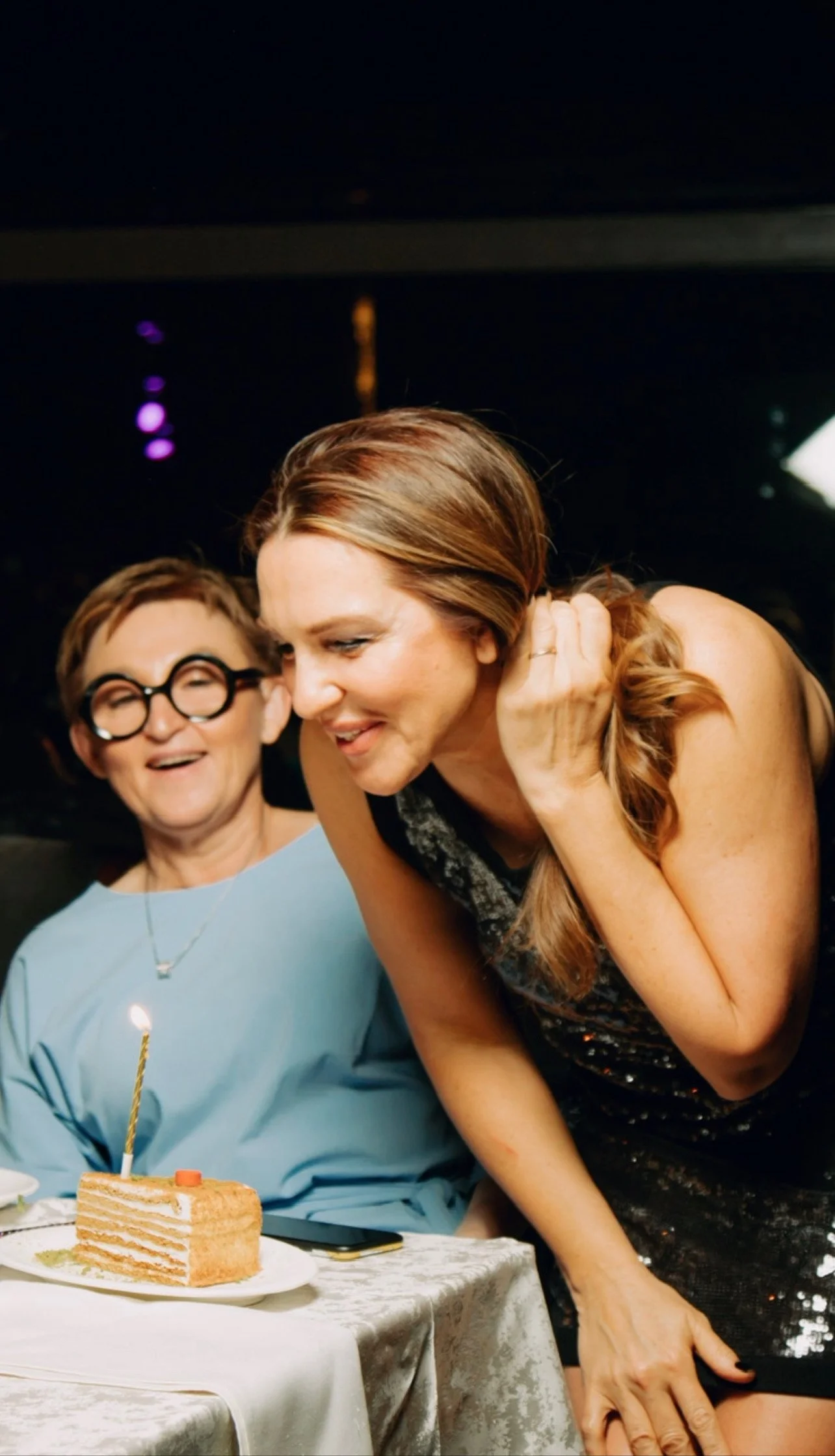 A birthday celebration with two women, one older with short hair and glasses, and one younger with long reddish hair, smiling and leaning over a table with a slice of cake with a lit candle and a single orange candy. They are in a dimly lit room with a dark background.