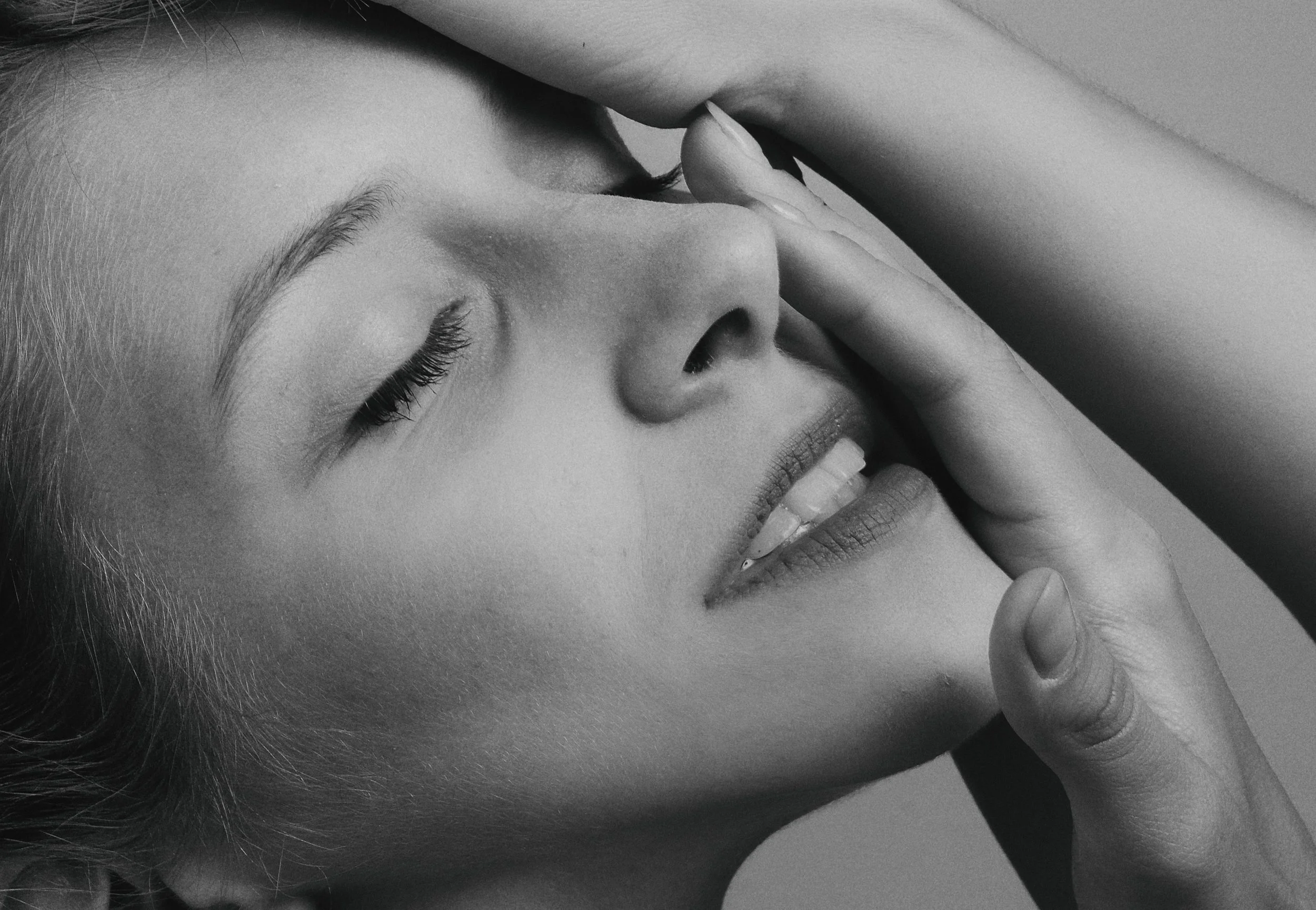 A close-up black and white photo of a woman with closed eyes, smiling slightly, holding her hand near her face with her finger touching her forehead.