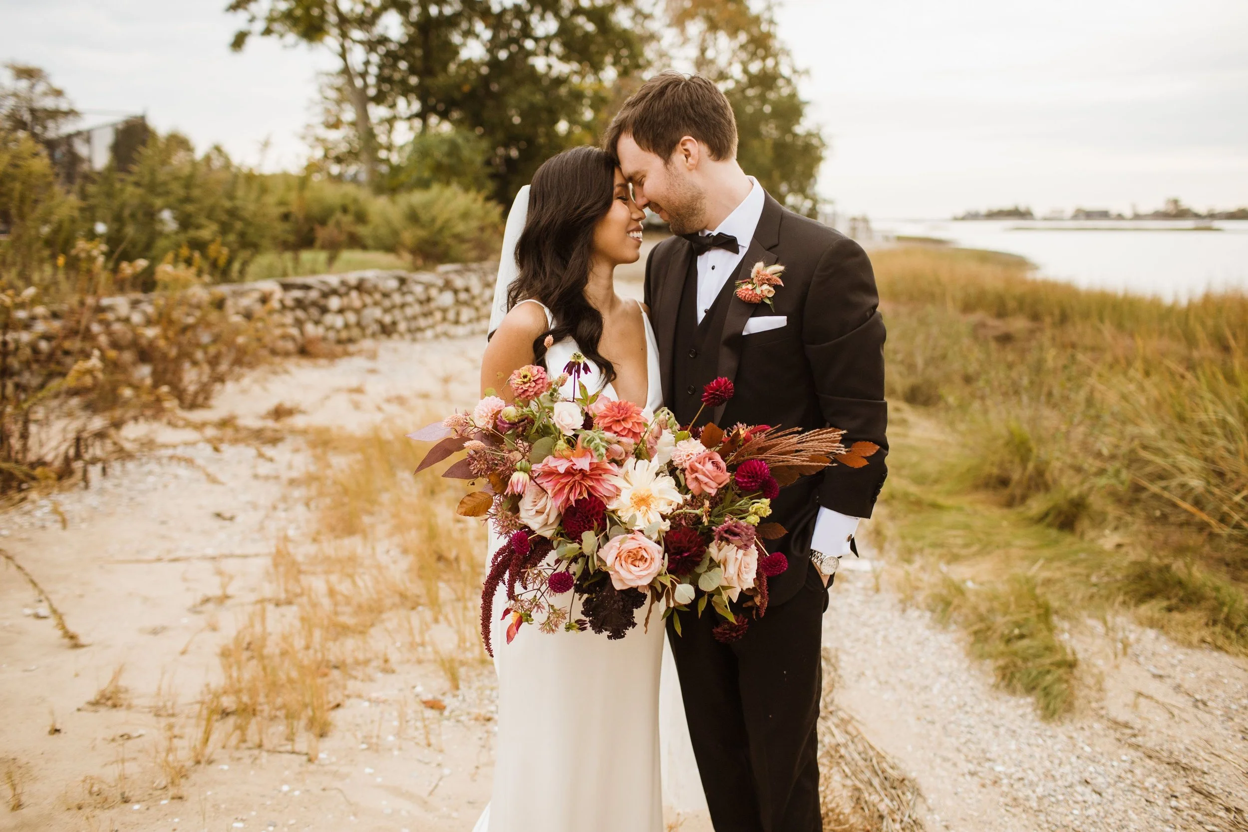 A bride and groom share a kiss outdoors during their wedding, with the bride holding a large bouquet of pink, red, and white flowers. The bride has long dark hair and is wearing a white wedding dress, while the groom is dressed in a black tuxedo with a bow tie and boutonniere. They stand on a sandy path near a stone wall, with trees and water in the background.
