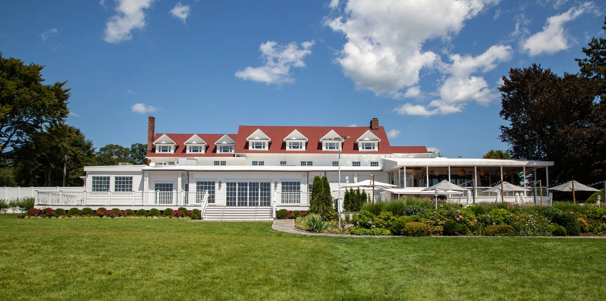A large white house with a red roof and multiple dormer windows, surrounded by a well-maintained lawn, colorful flowers, and trees under a partly cloudy sky.