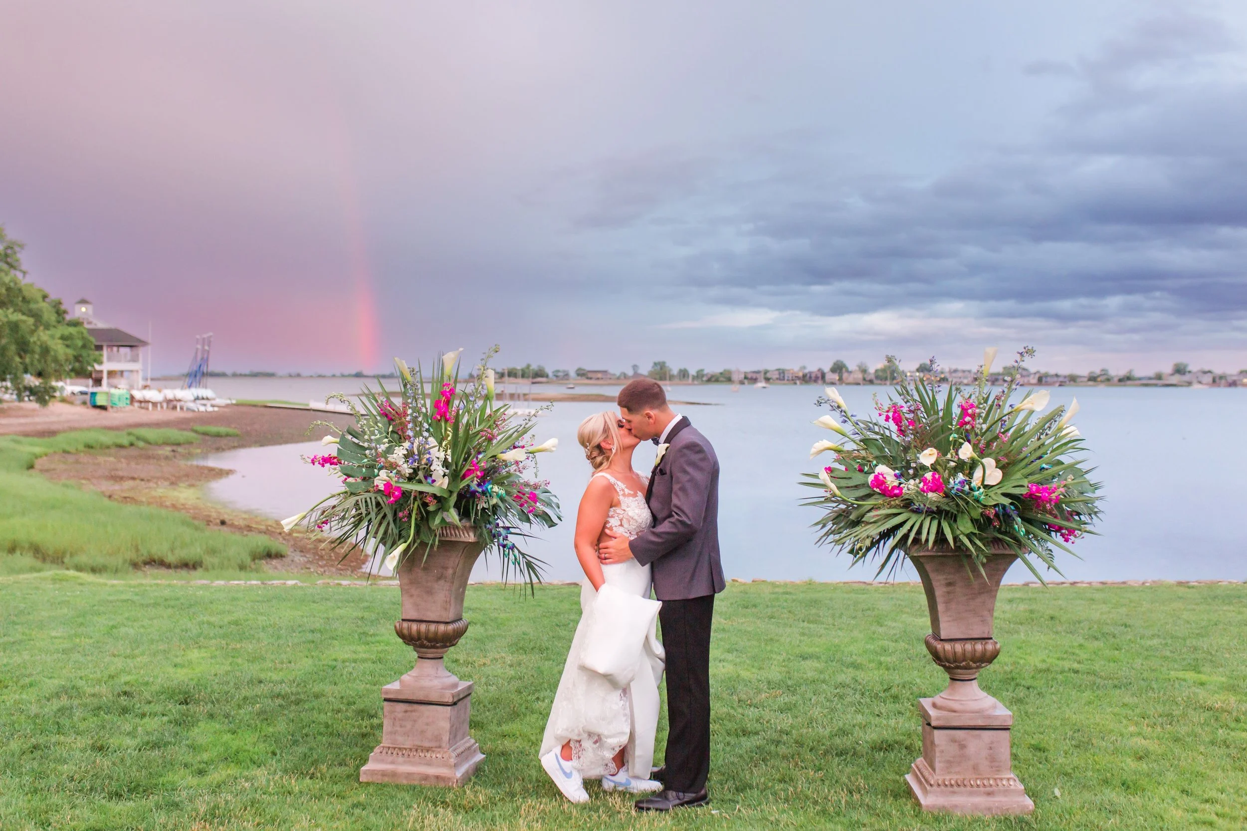 A bride and groom share a kiss during their outdoor wedding ceremony by a body of water. Two large flower arrangements on tall pots frame the couple, with a rainbow visible in the cloudy sky in the background.