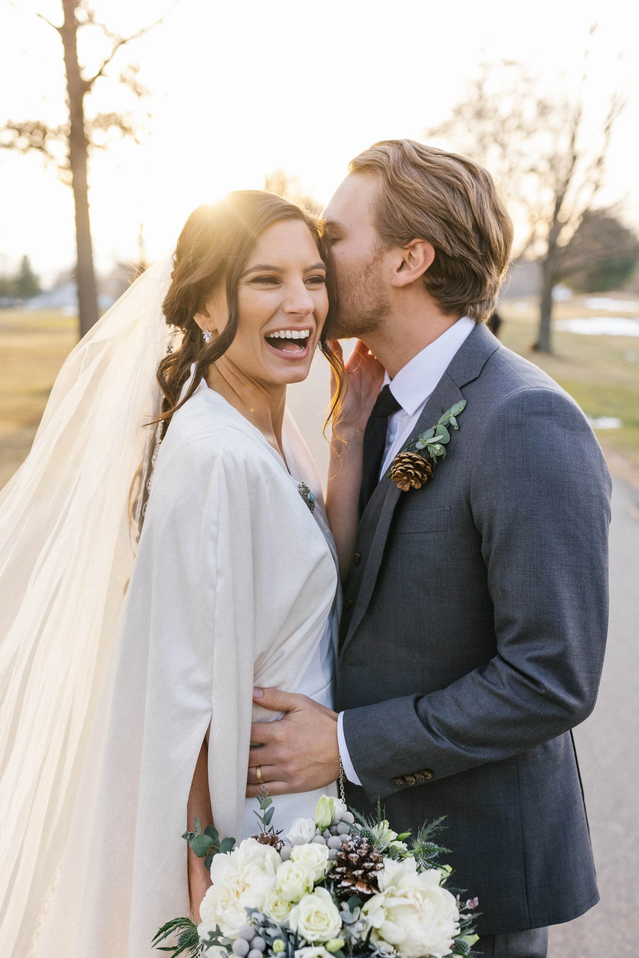 A bride and groom on their wedding day outdoor in the late afternoon or early evening with sunlight behind them. The bride has dark hair, is laughing, and holding a bouquet of white flowers. The groom is kissing her on the cheek, is wearing a gray suit, and has a pine cone boutonniere.