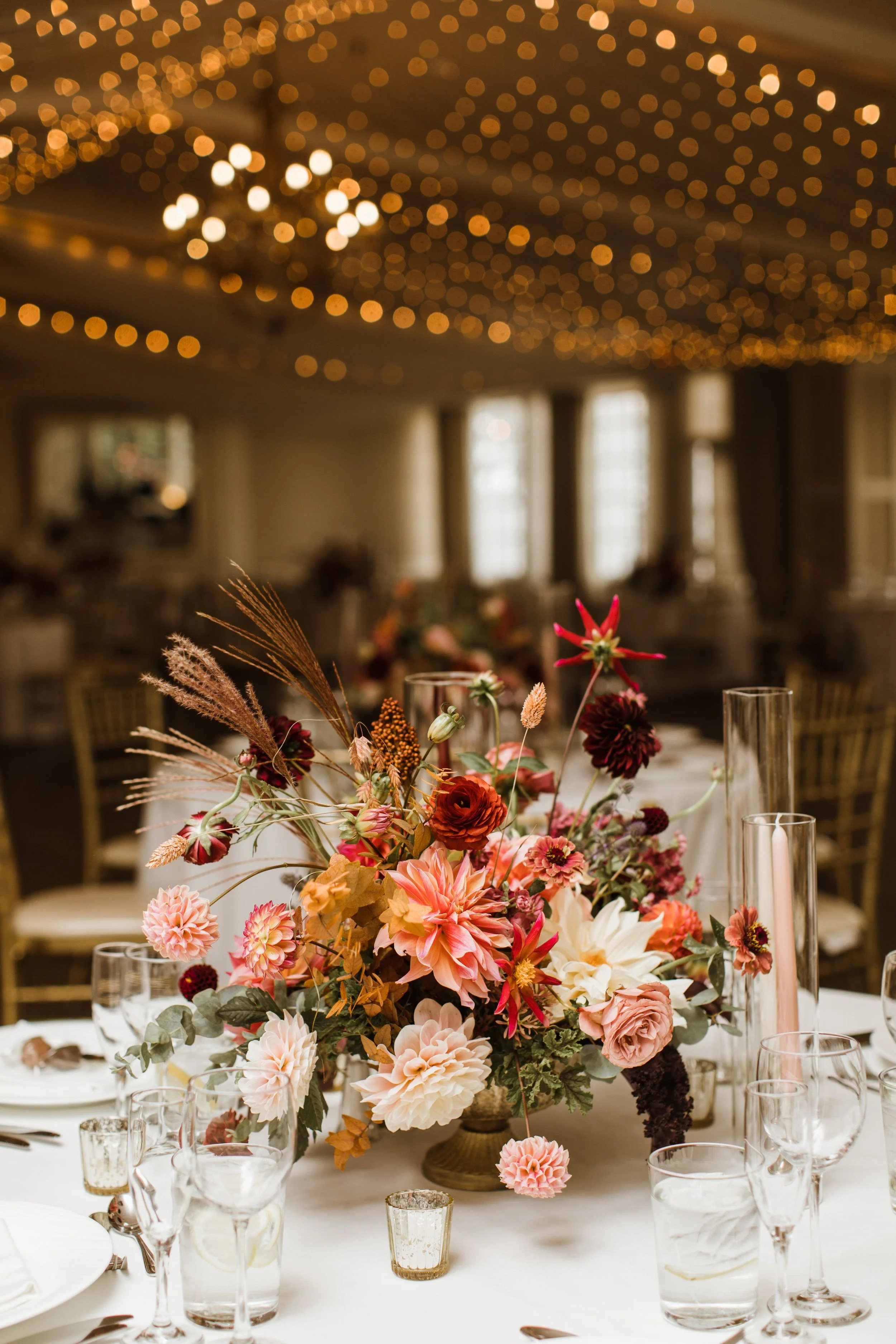 A table decorated with a large flower arrangement including pink, red, and white flowers with greenery, surrounded by glassware and candles in small holders. In the background, a room with soft lighting and bokeh string lights on the ceiling.