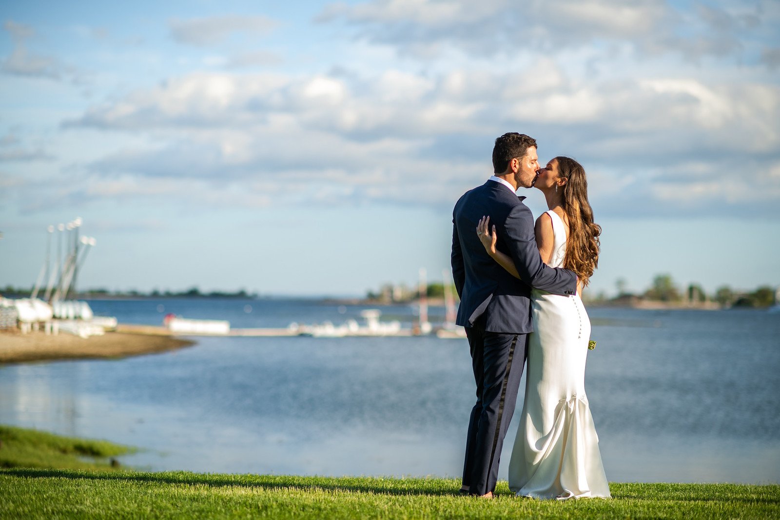 A bride and groom kissing outdoors near a body of water with a cloudy sky, green grass, and boats in the background.
