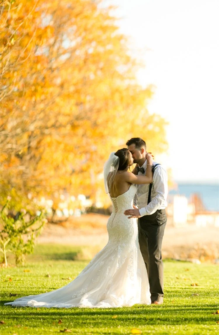 A bride and groom sharing a kiss outdoors on a sunny day, with autumn trees in the background.