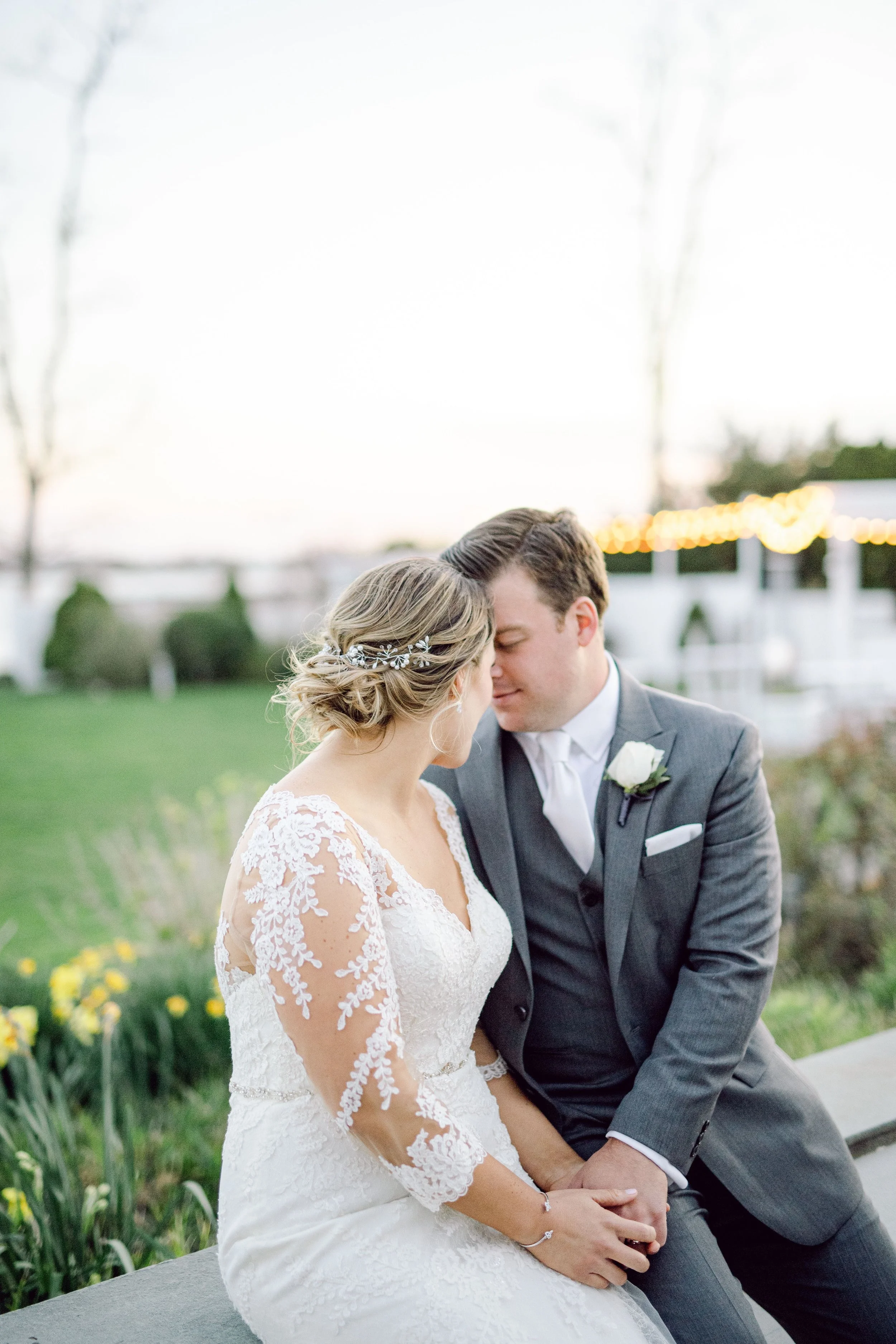 A bride and groom sit close together outdoors, touching foreheads with eyes closed, during sunset with string lights in the background.