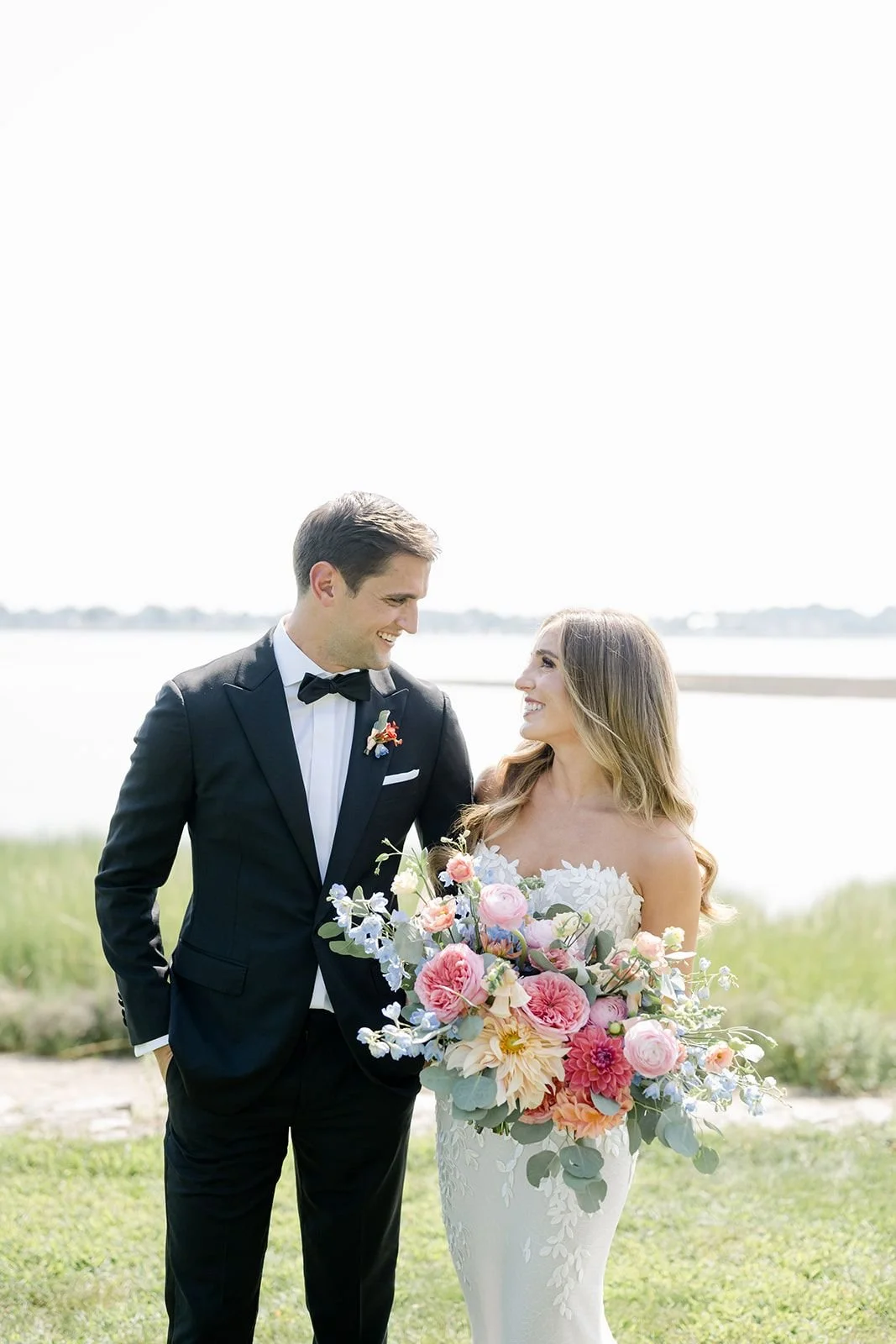 A bride and groom stand outdoors by a body of water, smiling at each other. The groom wears a black tuxedo with a bow tie, and the bride wears a white floral wedding gown, holding a large bouquet of pastel-colored flowers.