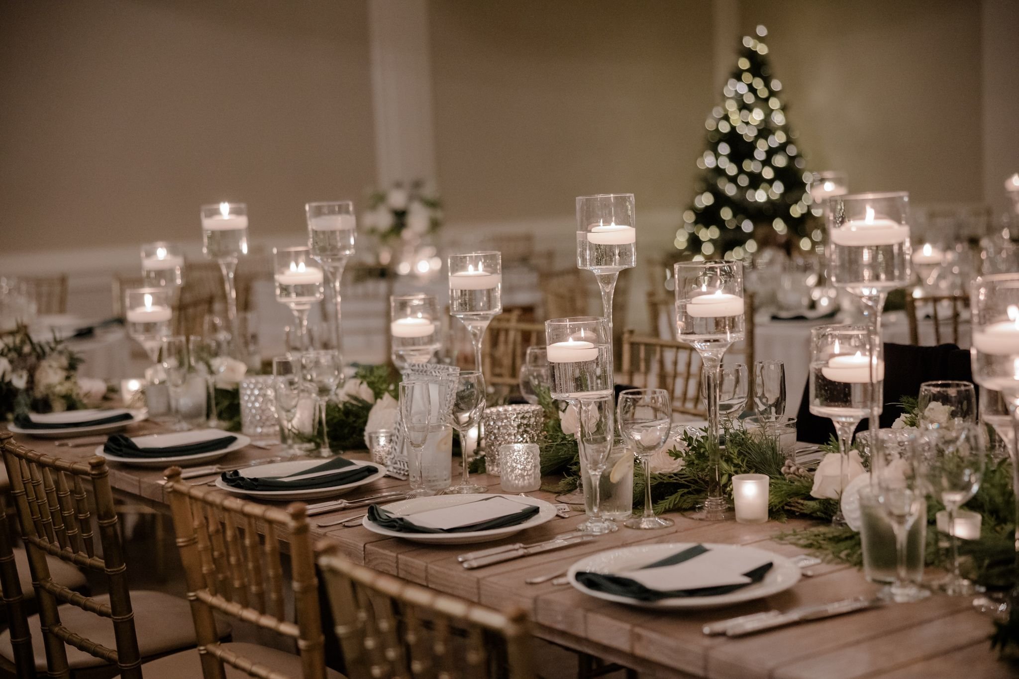 A decorated dining table with floating candle centerpieces and greenery, set for a holiday celebration with a Christmas tree in the background.