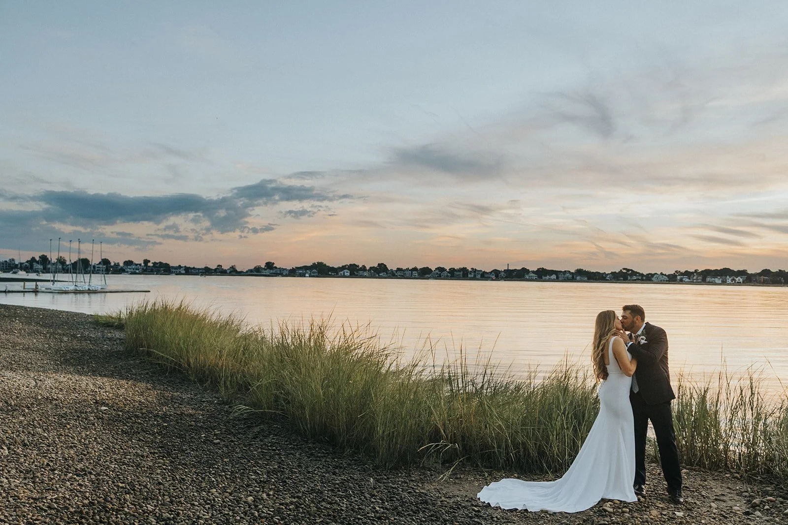 A bride and groom sharing a kiss on a rocky beach at sunset, with water and houses in the background.