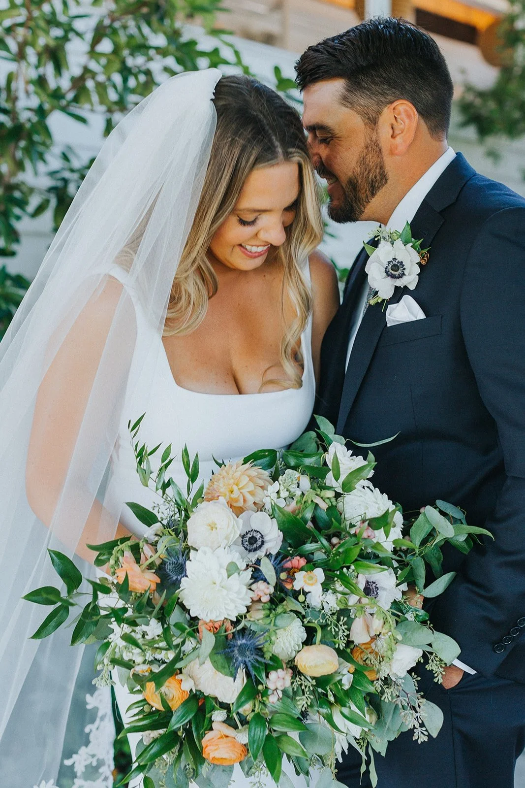 A bride and groom on their wedding day sharing a close, happy moment, with the bride holding a large bouquet of white, orange, and purple flowers.