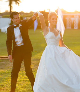 A newlywed couple dancing outdoors in the sunlight. The groom, in a tuxedo, holds the bride's hand as she twirls in a white wedding gown. They are smiling and enjoying their wedding celebration.
