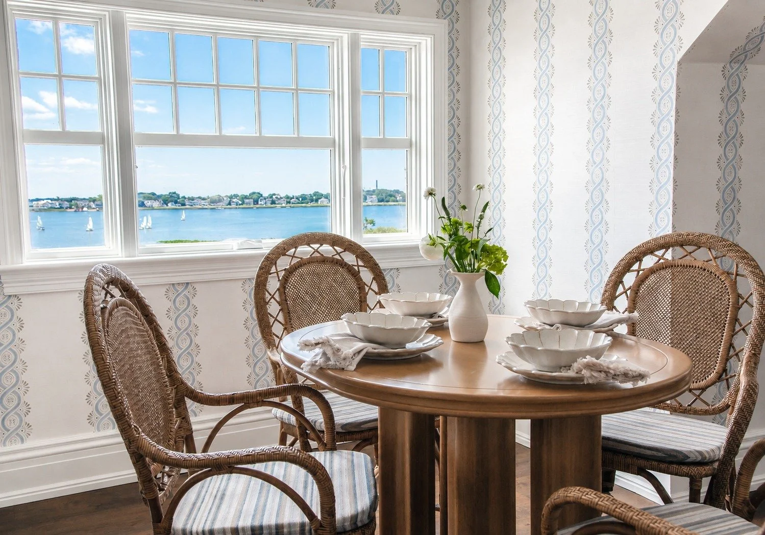 A dining room with a round wooden table set with white bowls and napkins, four wicker chairs with cushioned seats, a large window showing a view of a river with sailboats and houses in the distance, and a vase with white flowers on the window sill.