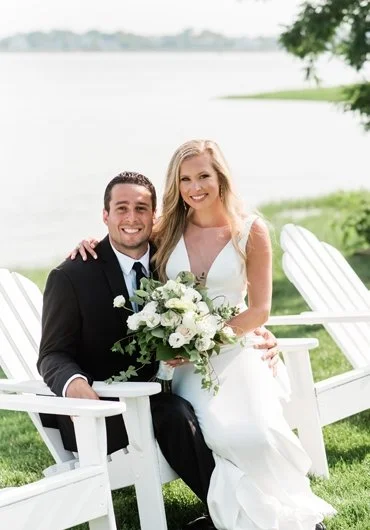 A bride and groom sitting on white garden chairs outdoors by a lake, smiling, with the bride holding a bouquet of white flowers.