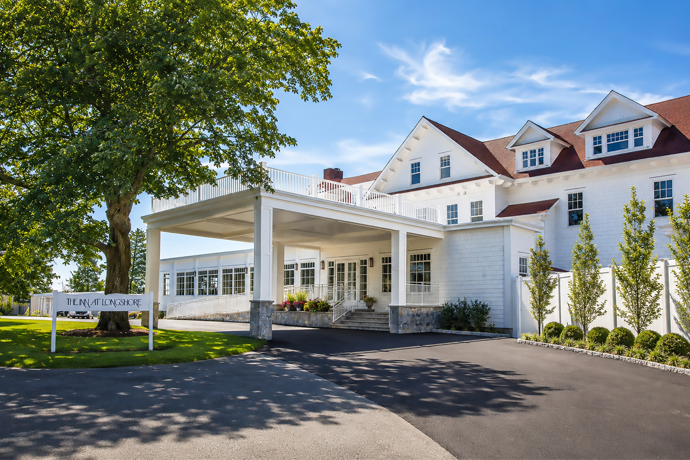 A large, white, multi-story house with a red roof and many windows, surrounded by trees and a well-maintained lawn. There is a sign that reads 'The Inn at Longshore' near the entrance.