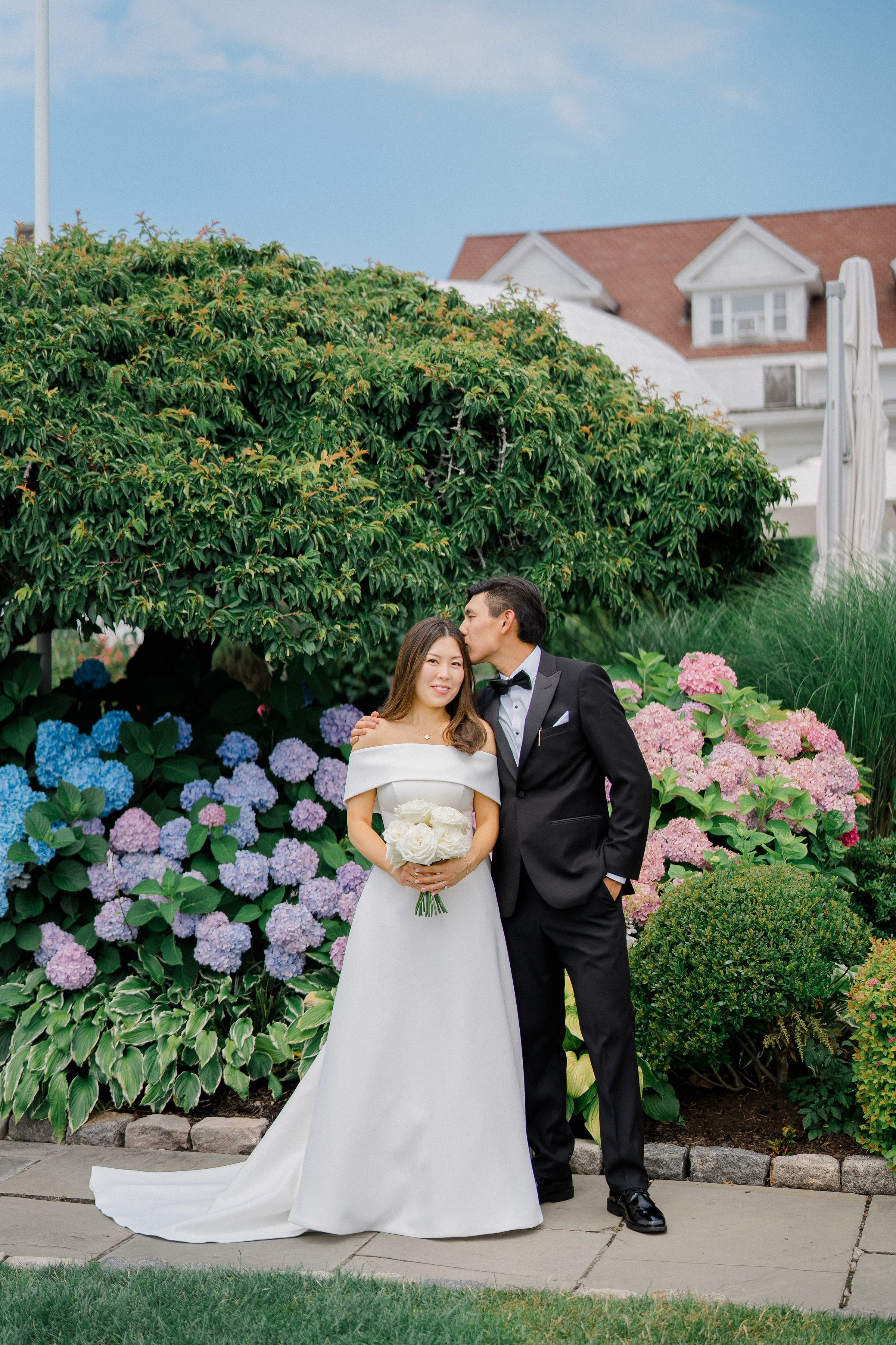 A newlywed couple standing in front of colorful hydrangea flowers and green bushes. The bride is wearing a white off-shoulder wedding gown and holding a bouquet of white roses, while the groom is wearing a black tuxedo and is kissing the bride on the forehead.