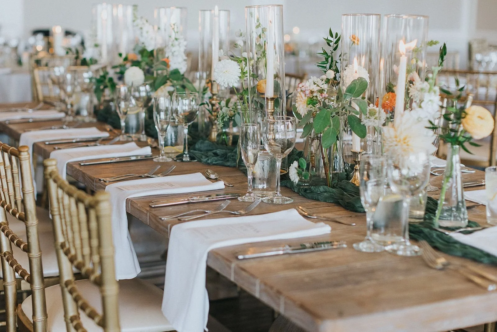 Elegant dining table decorated with white and orange flowers, green foliage, tall glass candle holders with lit white candles, and glassware set for a formal event, with gold chairs and a wooden table.