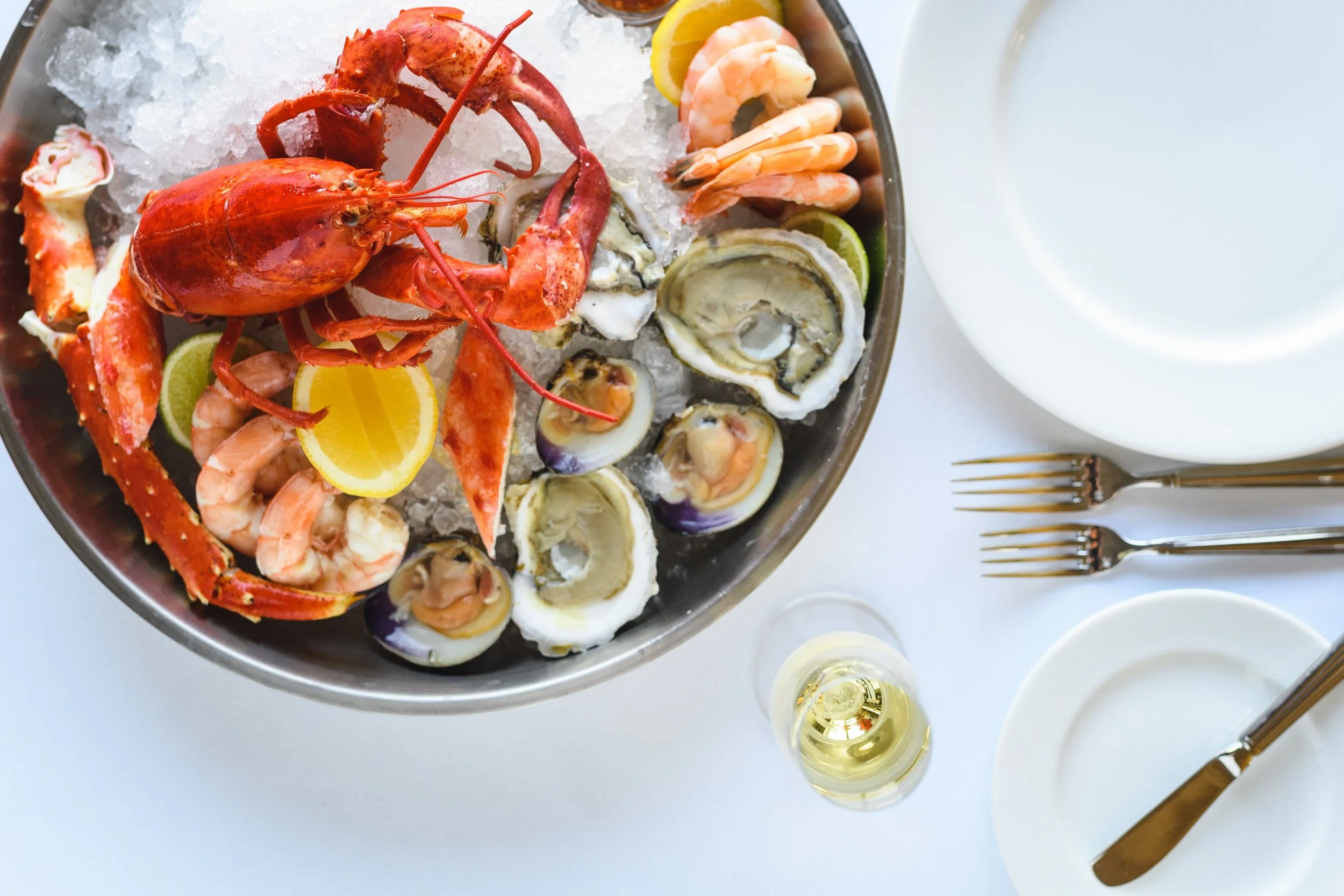 A seafood platter with lobster, shrimp, oysters on ice, and lemon wedges, served on a large silver tray. Surrounding the tray, there are white plates, golden forks, a glass of white wine, and a candle.