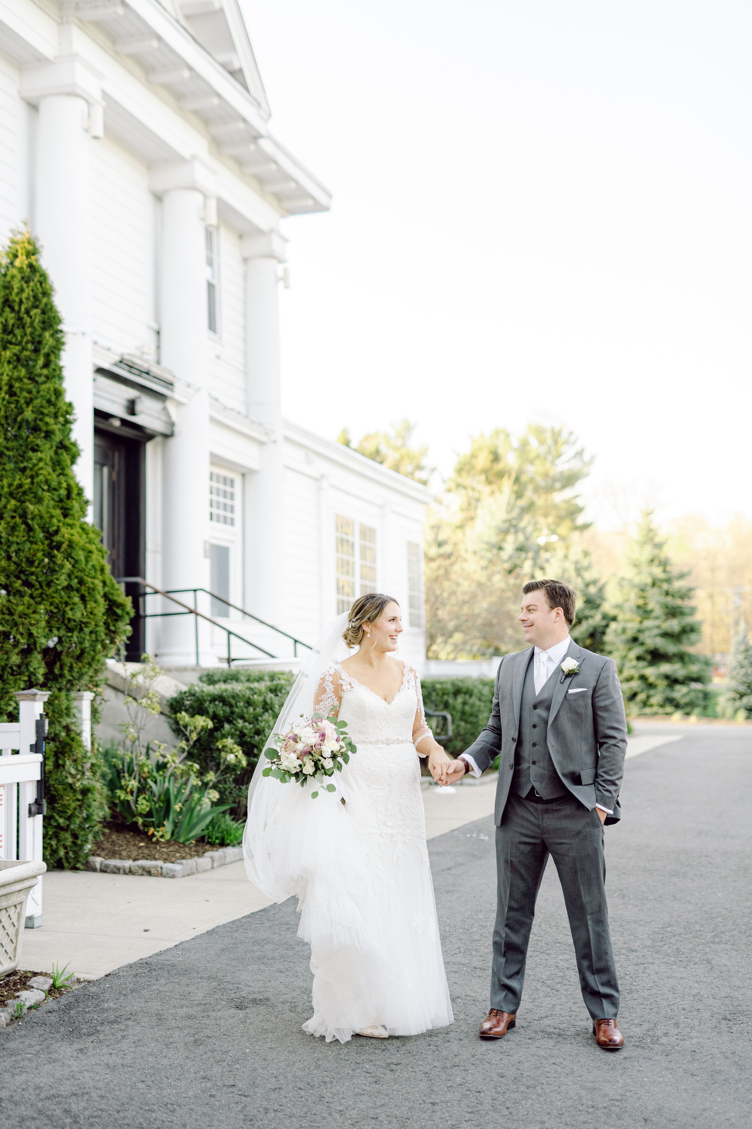 A bride and groom holding hands and smiling at each other outside a white building with columns, greenery, and trees in the background.