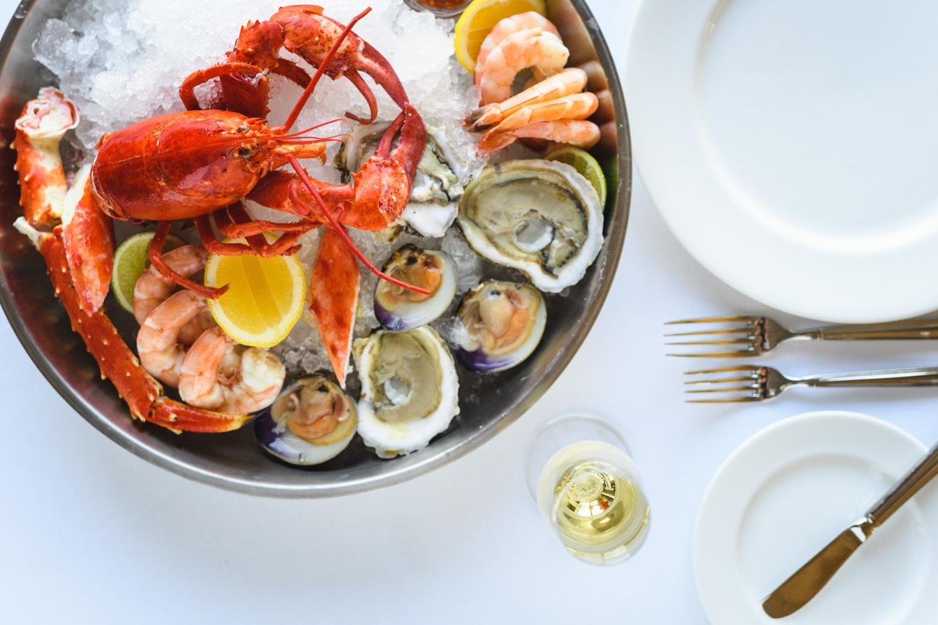 A seafood platter with lobster, crab, shrimp, oysters, and clams served on a bed of ice, with lemon and lime wedges, a glass of white wine, and tableware set for dining.