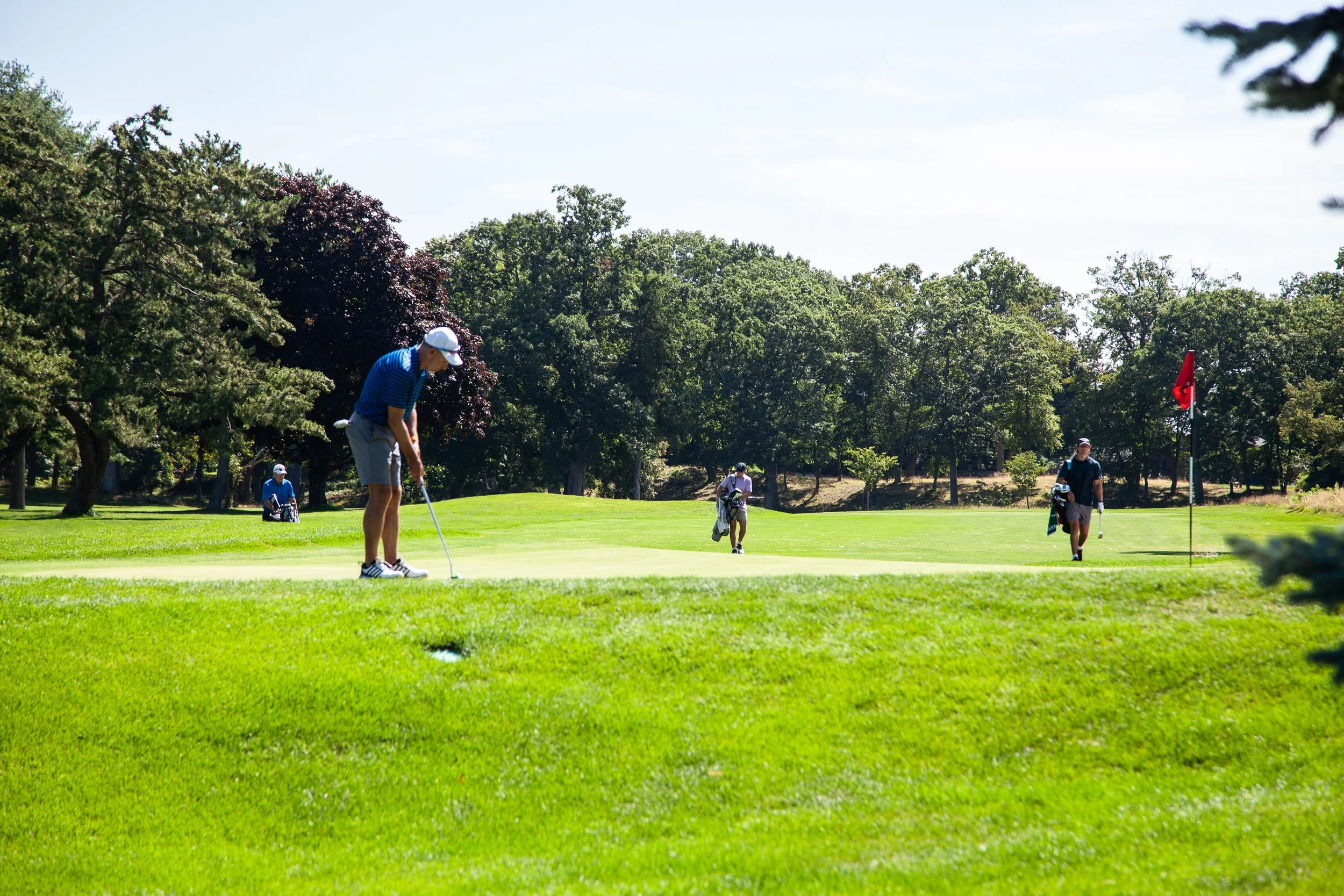 People playing golf on a sunny day at the golf course, with trees and a red flag on the green.