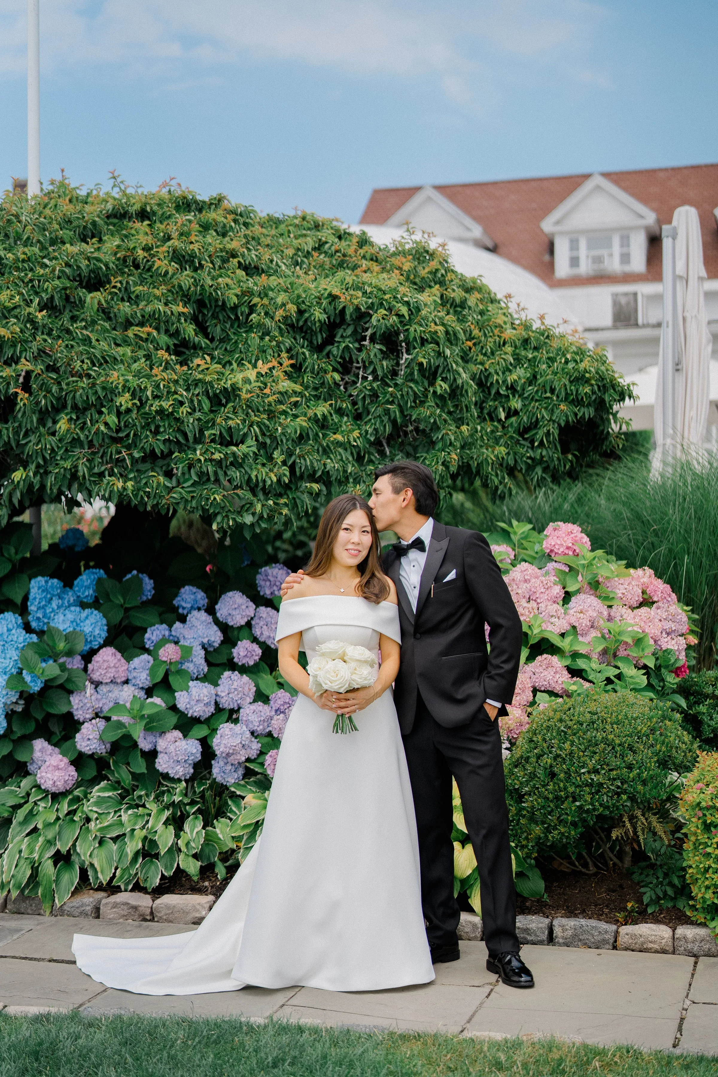 A bride and groom standing together in front of lush green bushes and colorful hydrangeas, with the groom kissing the bride on her forehead. She is holding a bouquet of white roses, and they are dressed in wedding attire. A house with a red roof and 