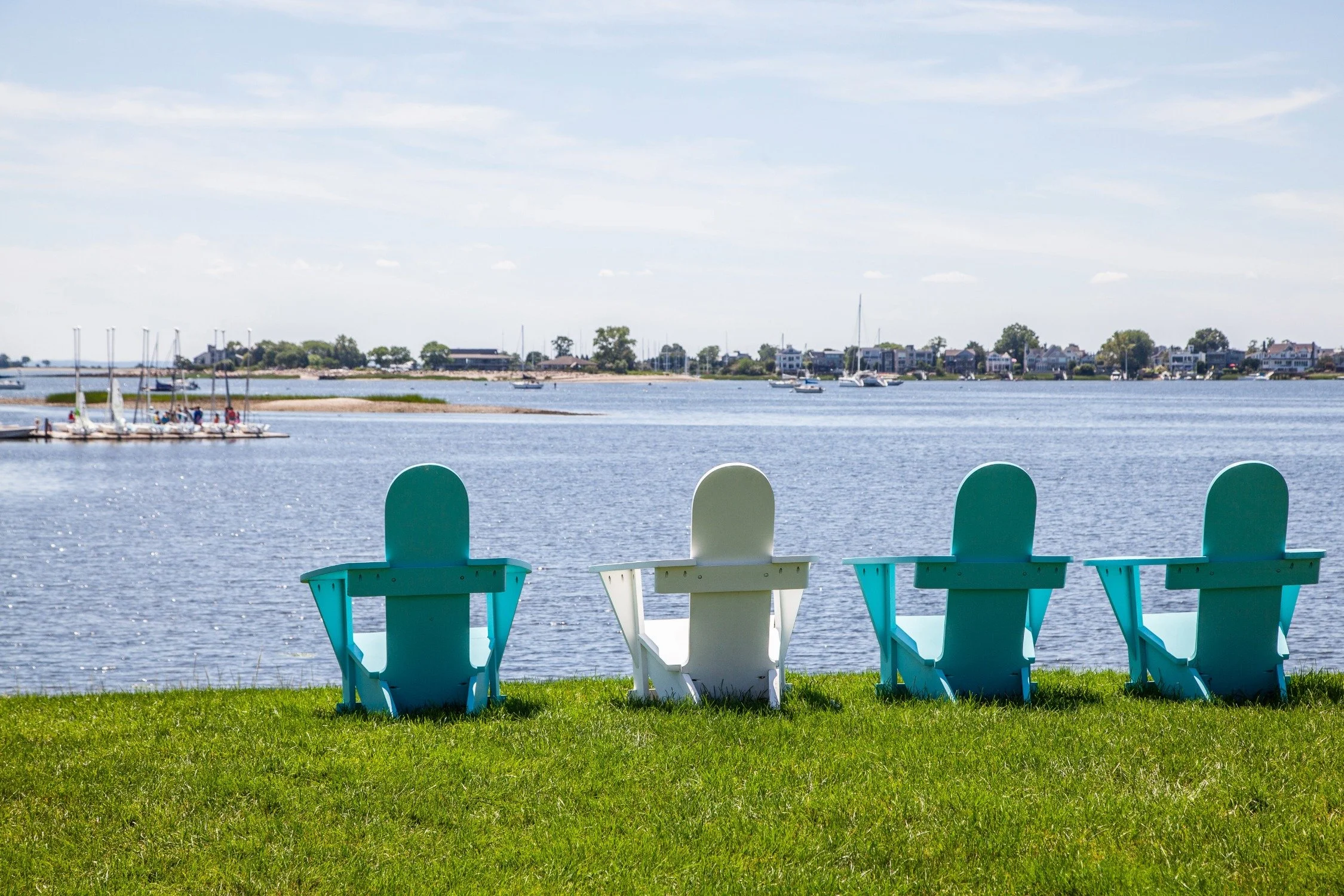 Four colorful Adirondack chairs facing a body of water with sailboats and houses in the background on a sunny day.