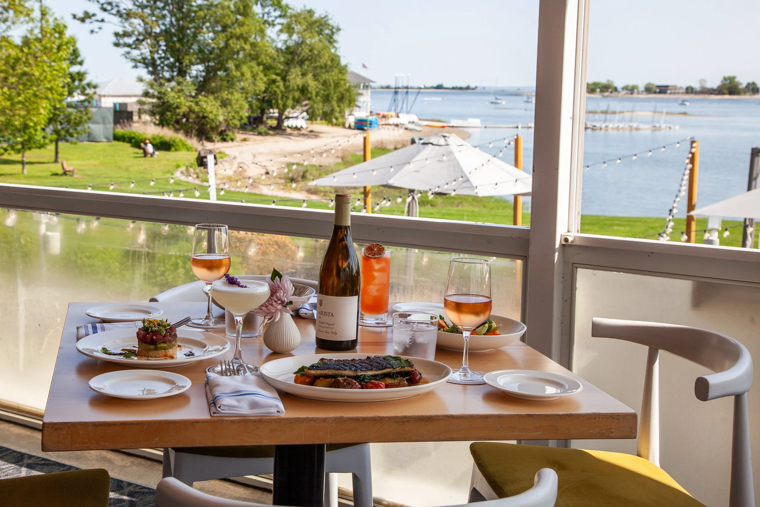 A dining table set with various dishes and drinks, overlooking a scenic waterfront view with trees, boats, and a patio umbrella outside the window.