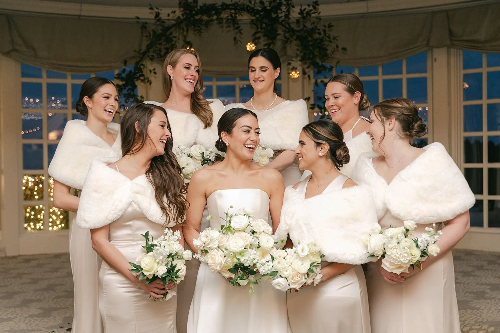 A group of women at a wedding, with the bride in the center holding a bouquet of white roses. The other women, likely bridesmaids, are gathered around her, all smiling and dressed in elegant white attire with some wearing white shawls. They are indoors with a decorated background and large windows.