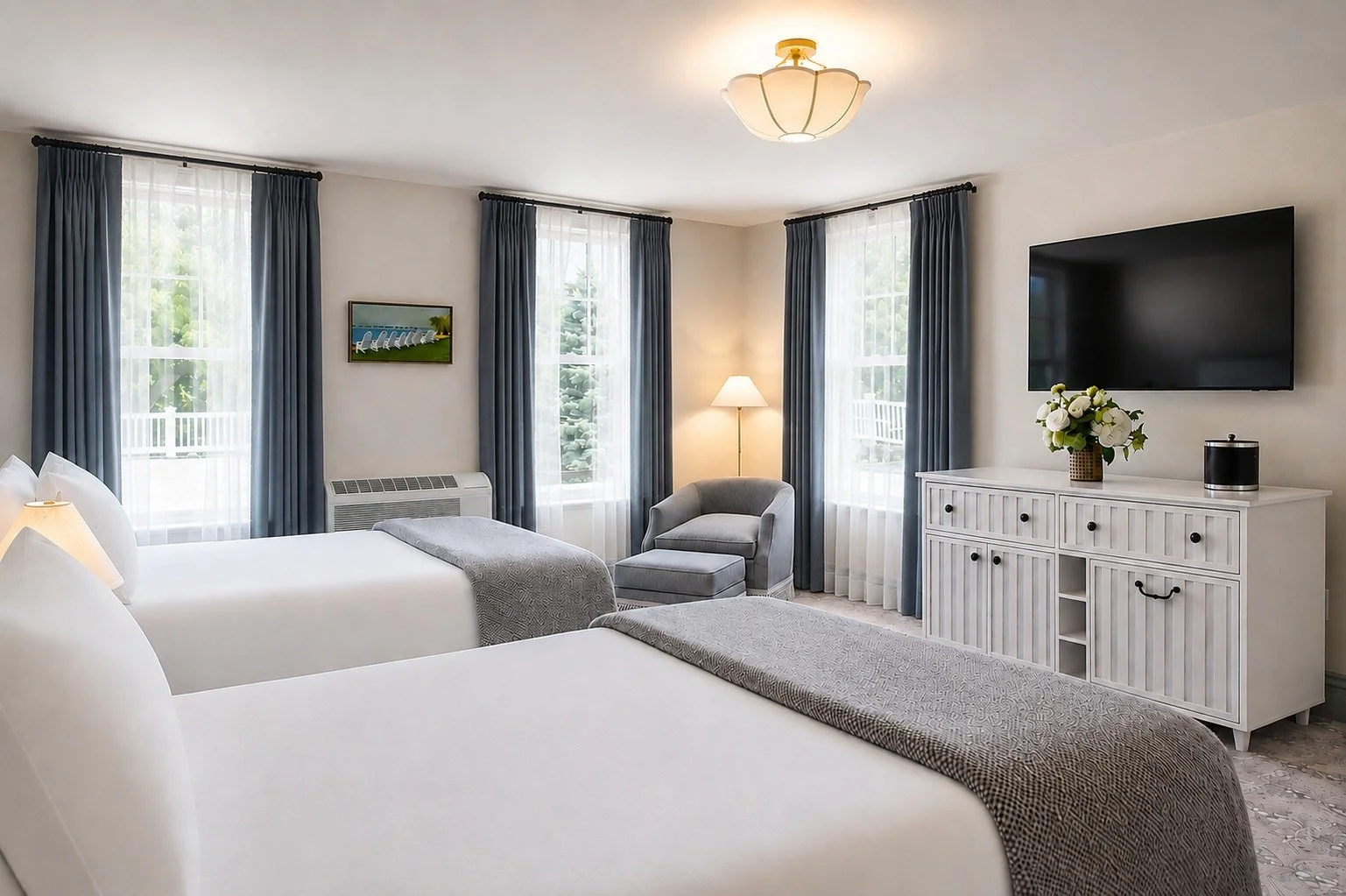 A hotel room with two queen beds, a grey armchair with ottoman, a television mounted on the wall, a white cabinet with a floral arrangement, and three large windows with blue curtains.