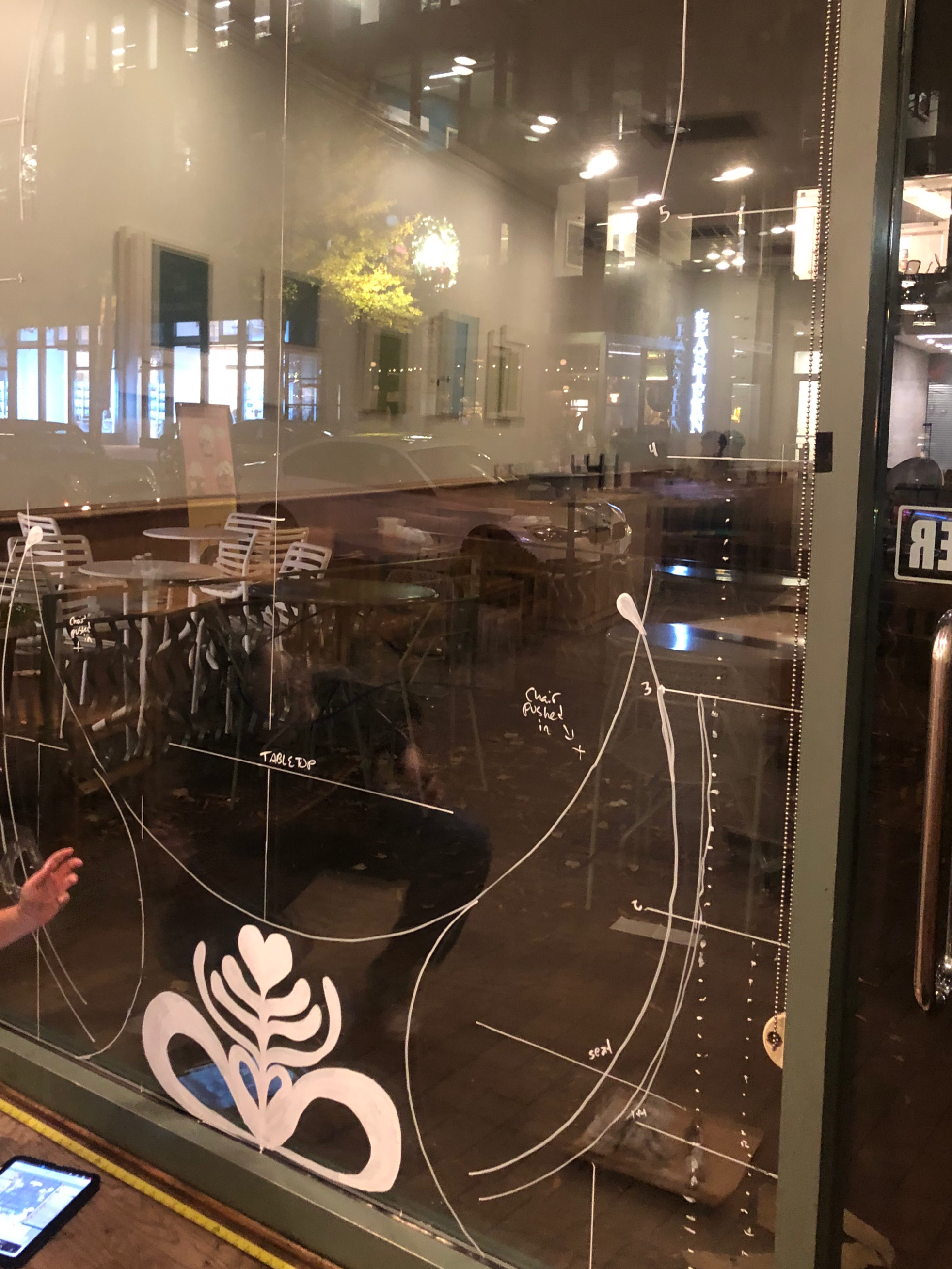 Nighttime view of a cafe through a glass window decorated with white painted floral and heart designs, including some handwritten notes.