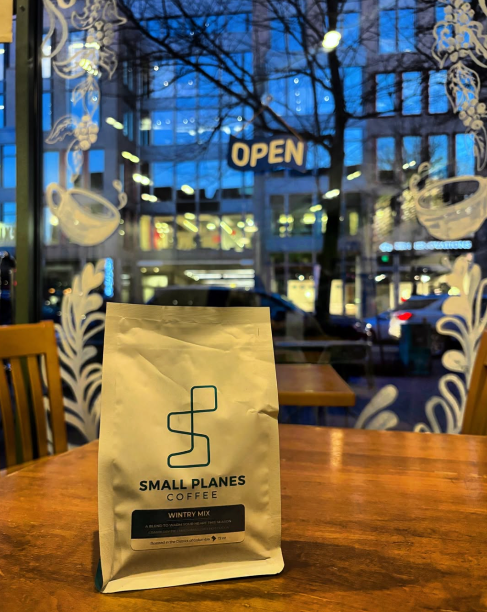 A bag of Small Planes Coffee Wintery Mix on a wooden table inside a coffee shop. Outside the window, a city street with parked cars and buildings, and a blue twilight sky. An "OPEN" sign hangs in the window.