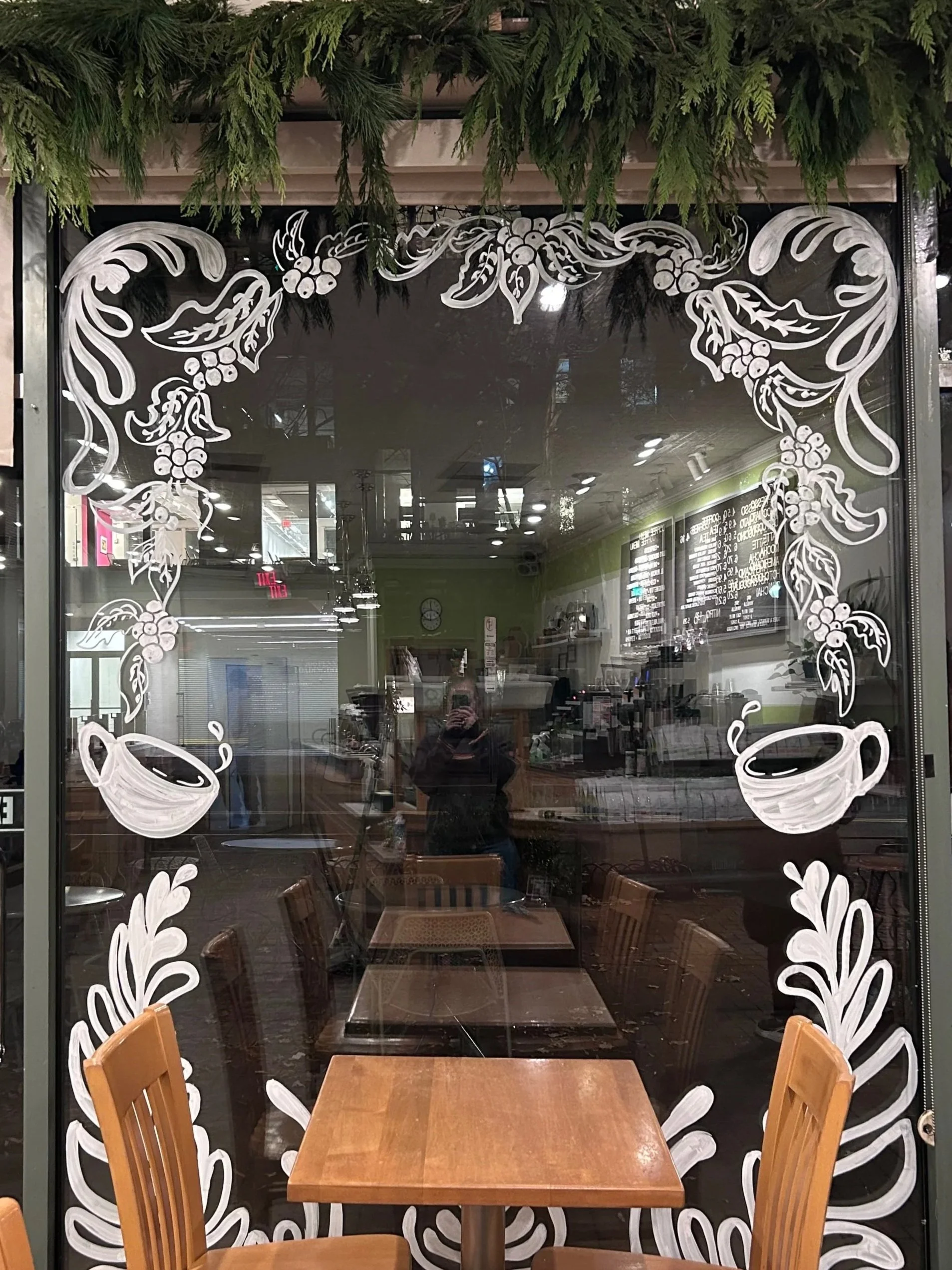 A view of a restaurant interior through a window decorated with white floral and coffee cup illustrations. Inside, there are wooden tables and chairs, with some overhead lighting and a menu board on the wall.