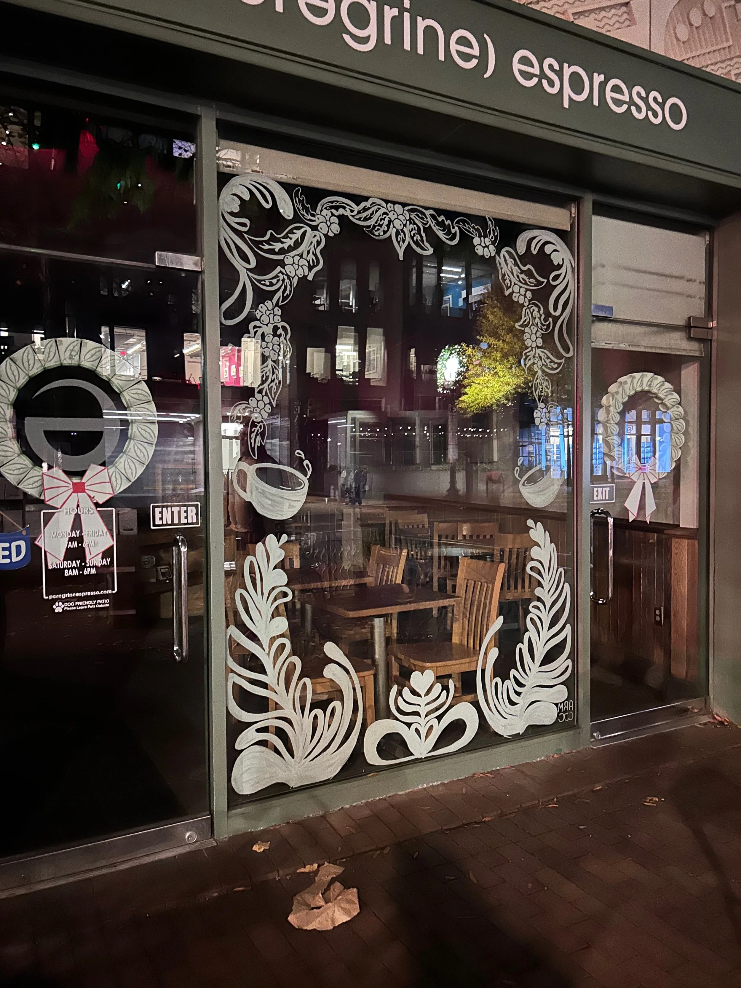 Store window decorated with white wreath and mistletoe designs, with wooden chairs visible inside, and signs indicating hours and entry.