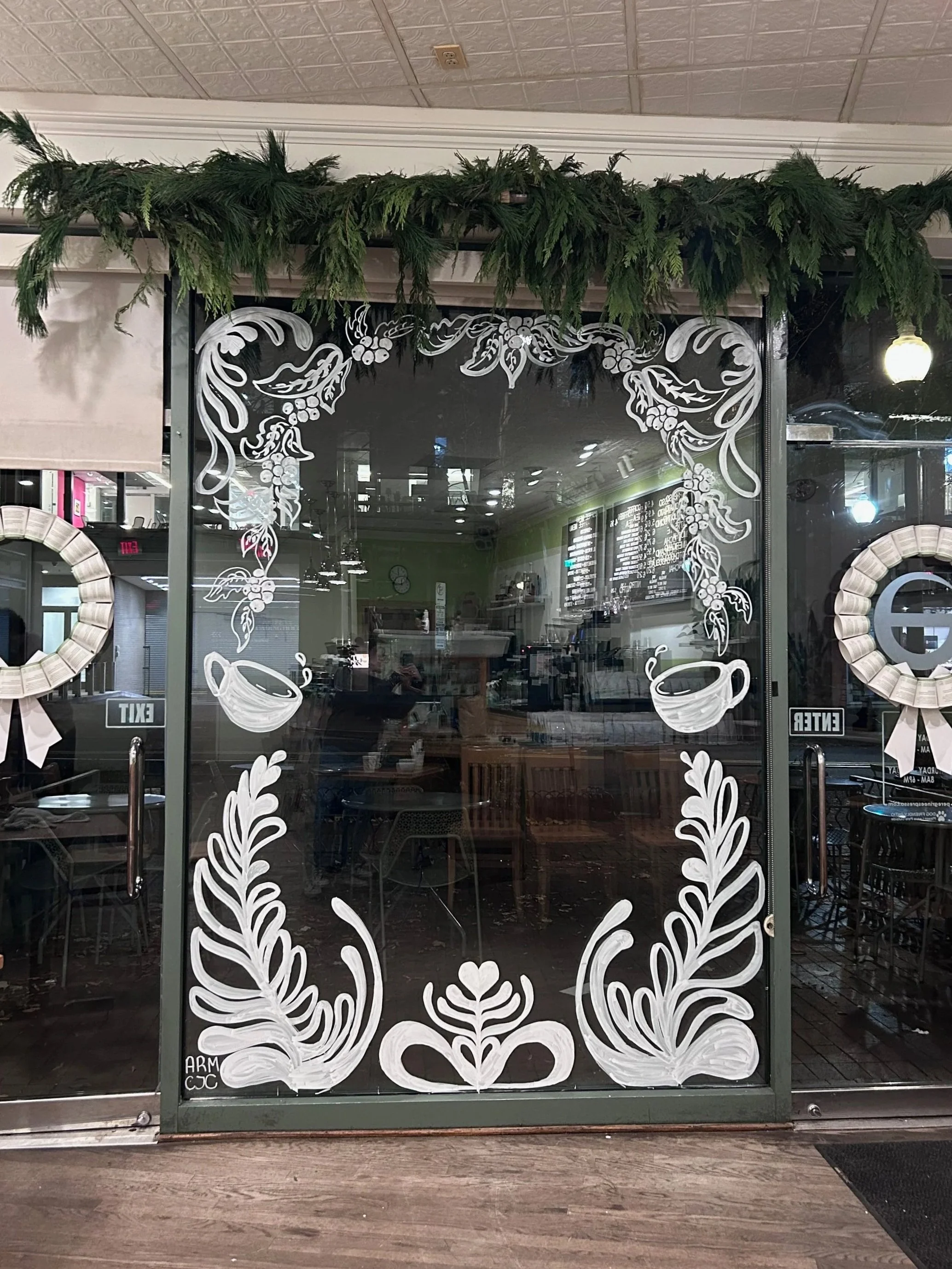 Decorative storefront window with white floral and coffee cup illustrations and greenery garland above the frame, inside a coffee shop.