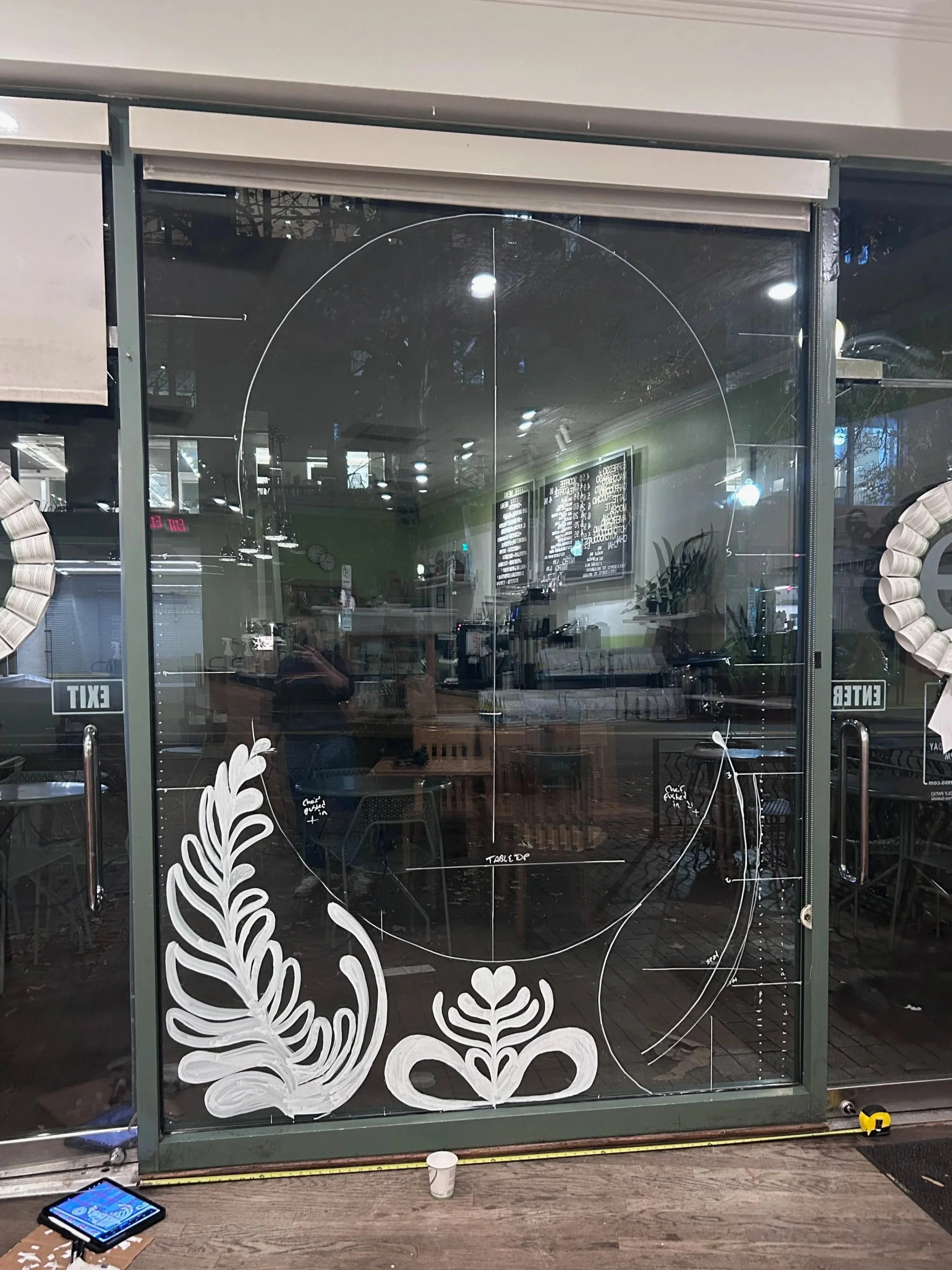 Glass door with white floral designs and handwriting indicating measurements, on a wooden floor inside a building, with reflections of the interior visible in the glass.