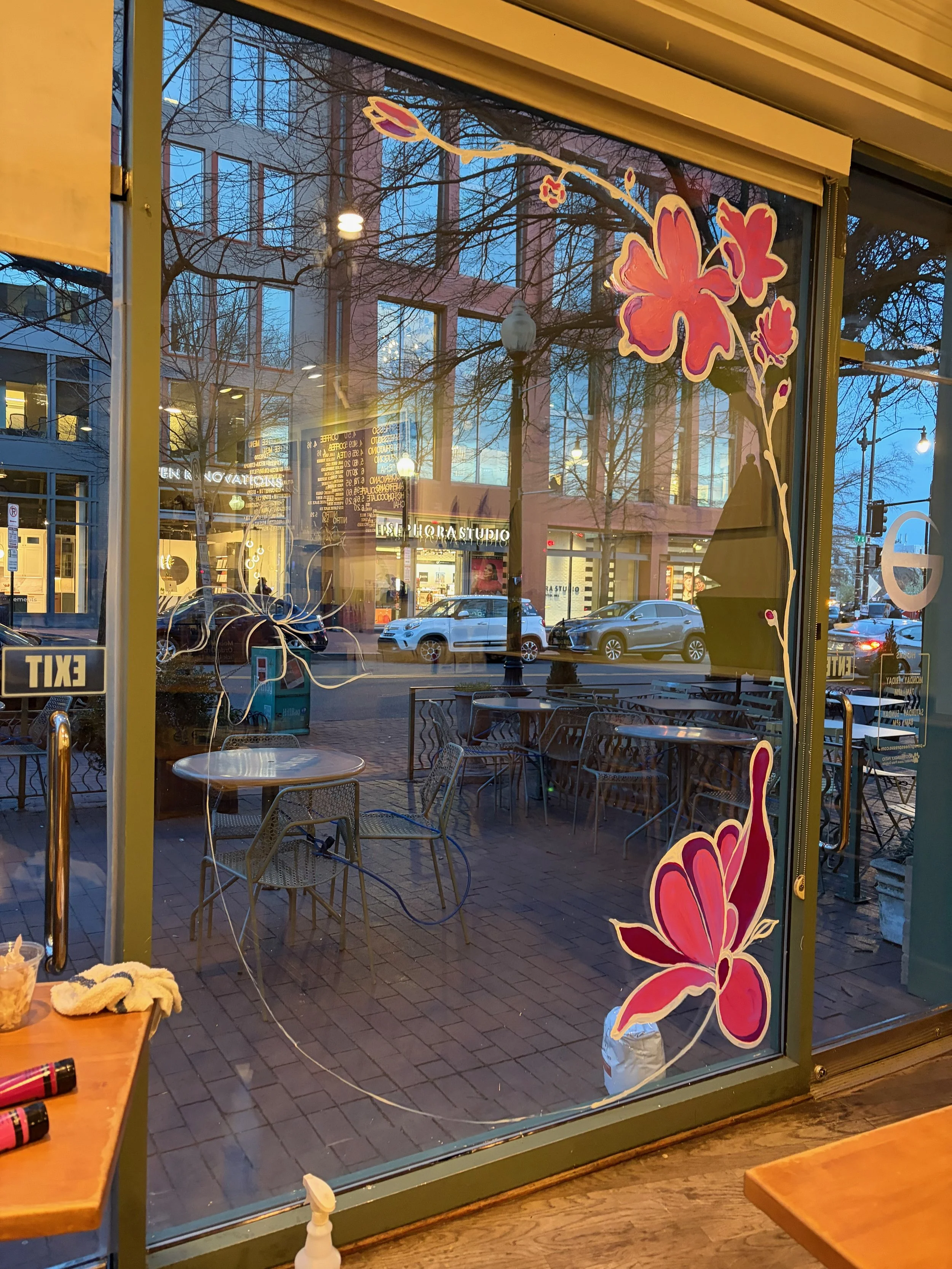 A street scene viewed through a restaurant window morning or evening, with reflections of buildings, trees, and streetlights. The window has floral decorations in pink and red.