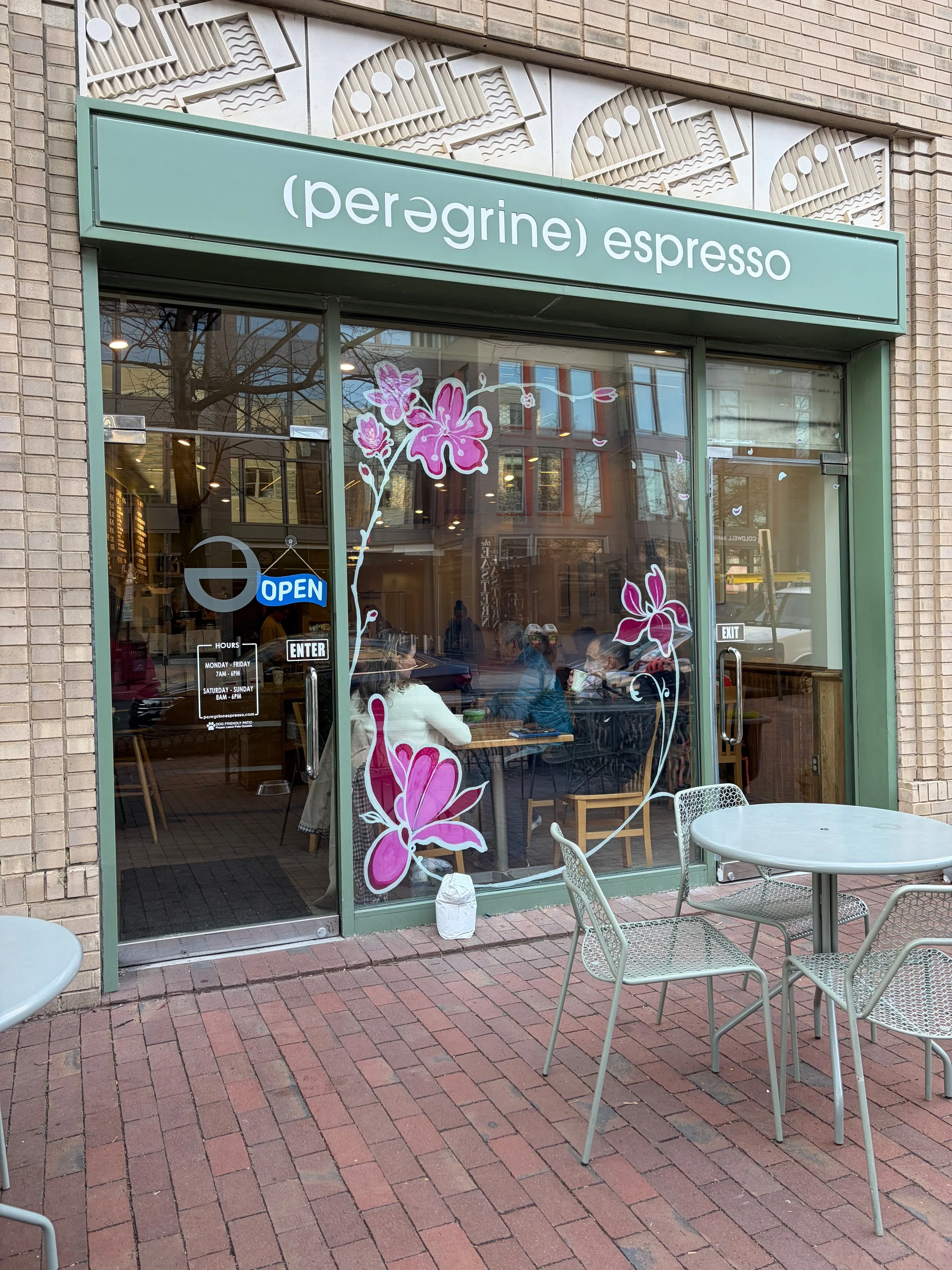 The exterior of a coffee shop named '(peregine) espresso' with a glass door and window decorated with pink flowers. Outside, there are empty tables and chairs on a brick sidewalk. Inside, customers are sitting and dining.