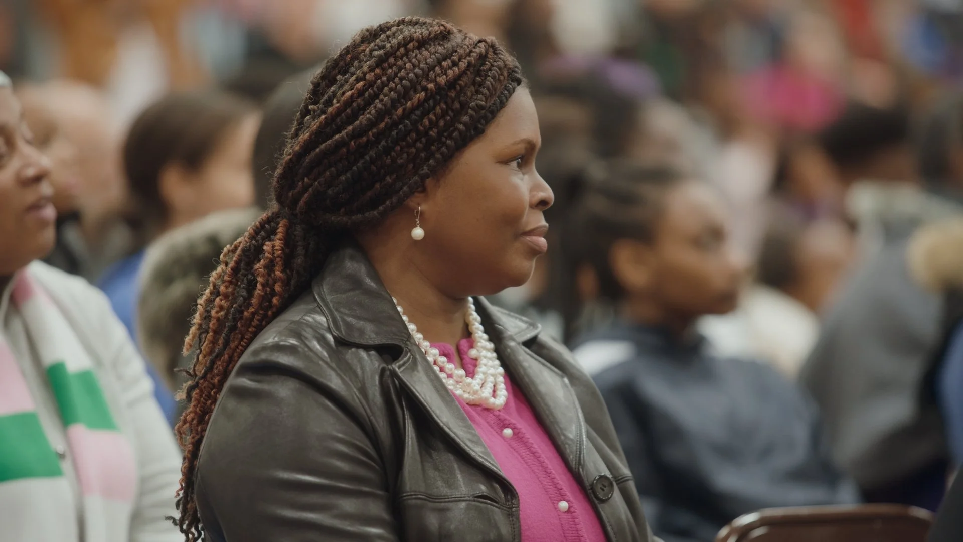 A woman with braided hair, pearl earrings, and a pearl necklace, wearing a leather jacket and pink top, attending an indoor event with other people seated around her.
