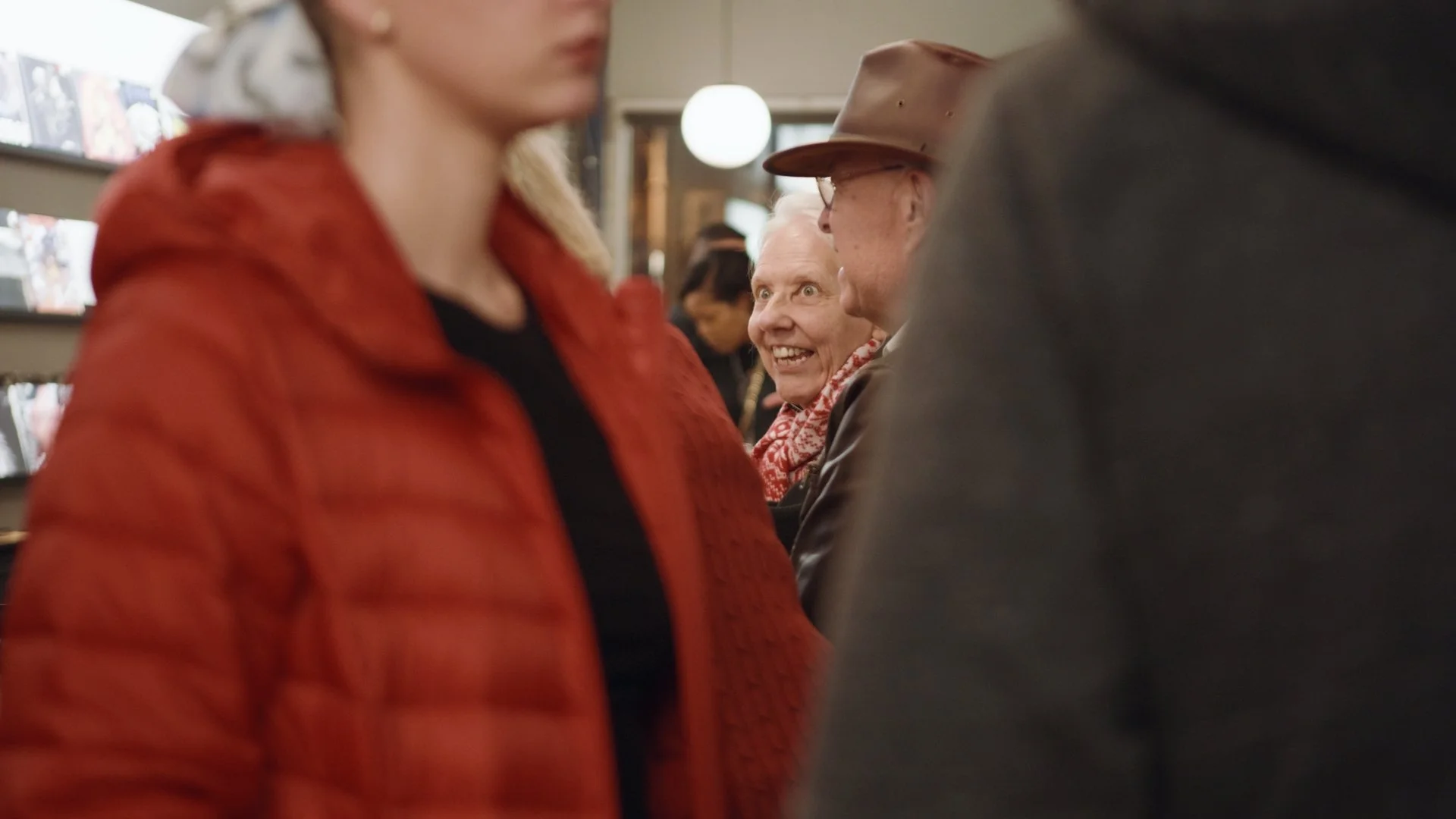 A group of people standing in line indoors, with a smiling elderly woman in the background. The focus is on the woman who is wearing a red shirt and red patterned scarf, and she is facing the camera. The other people are partially visible, wearing da