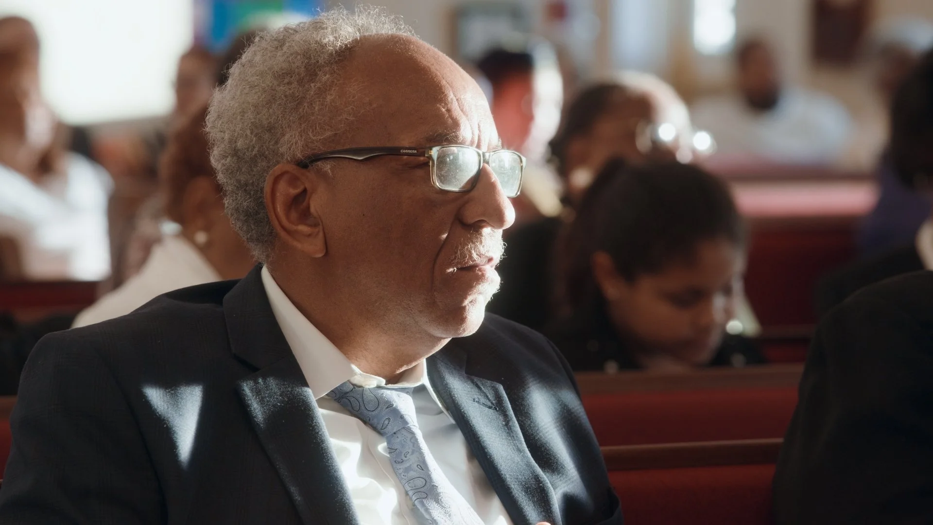 An elderly man with gray hair, glasses, and a white mustache is sitting in a church or auditorium wearing a suit and tie. He appears to be attending a formal event with other people in the background.