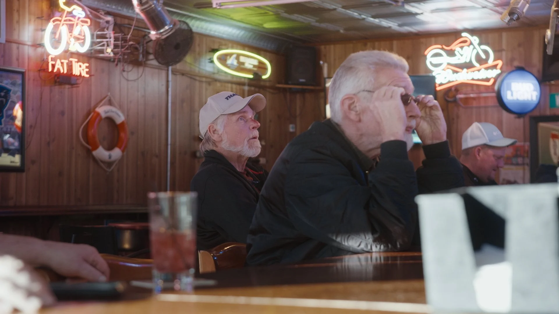 Three older men sitting at a table in a bar or restaurant with wood-paneled walls and neon signs, one adjusting sunglasses, another wearing a baseball cap, and the third looking straight ahead.
