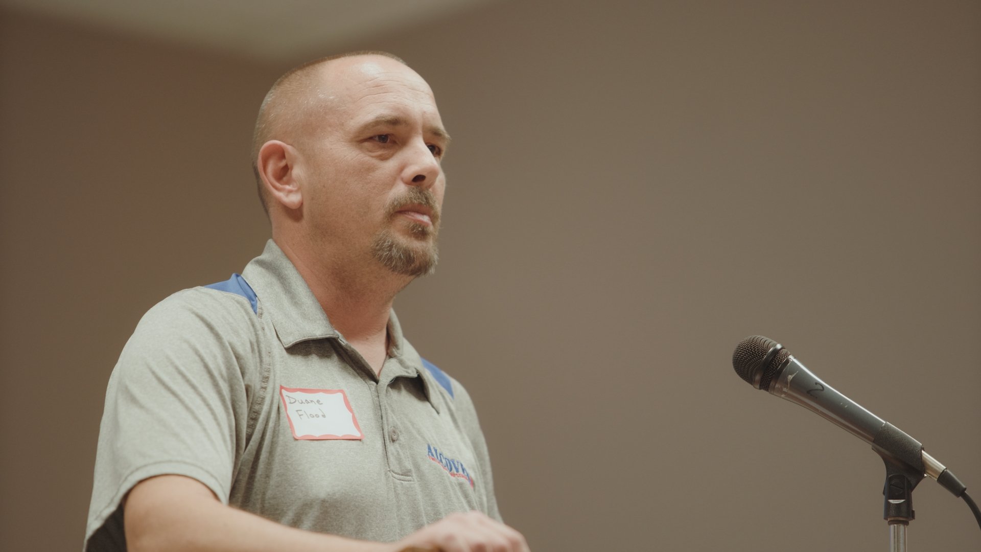 A man with a shaved head and goatee speaking into a microphone, wearing a gray uniform with a name tag that reads 'Duane Flood'.