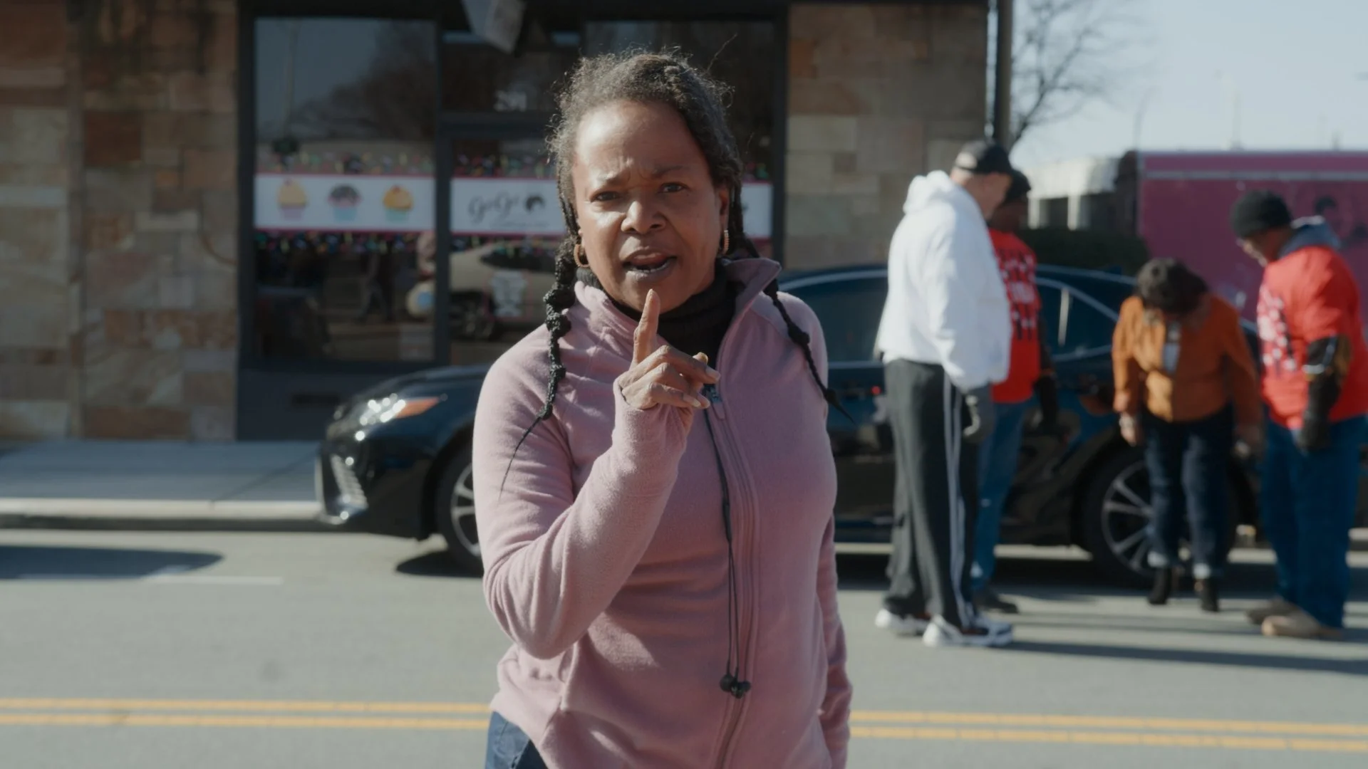 A woman with braided hair and earrings, pointing her finger and speaking outdoors in front of a group of people near a parked car and storefront.
